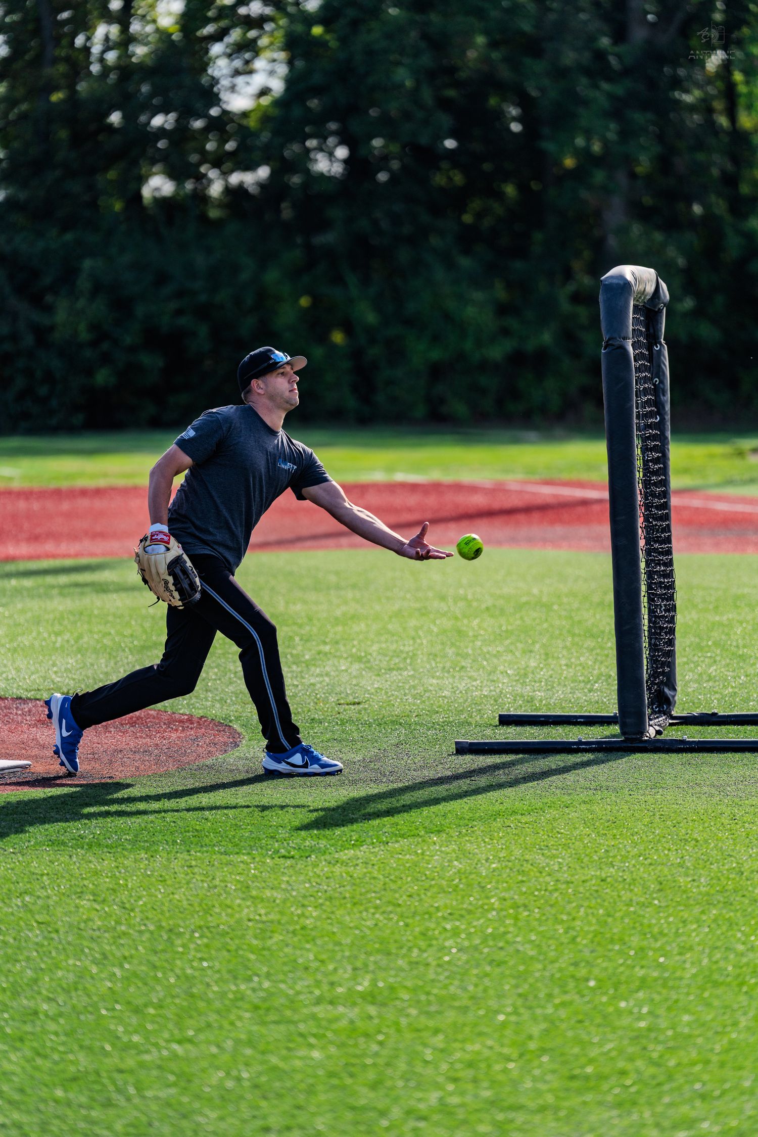 Male softball player in black uniform releasing a pitch on a green turf field with trees in background.
