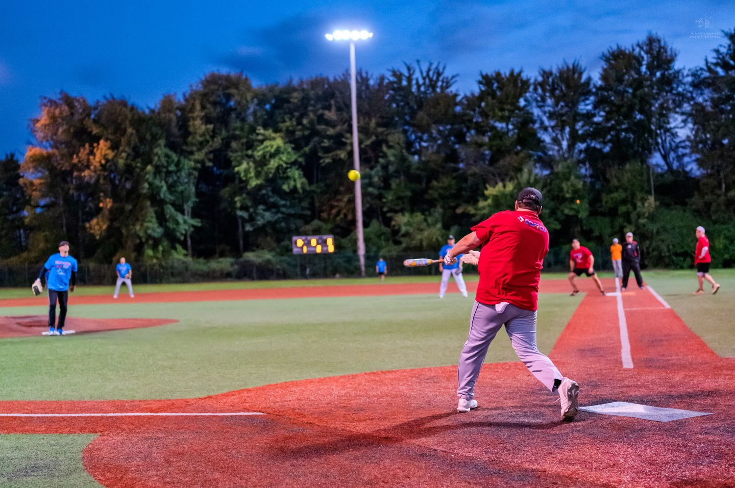 A batter swings at a pitch during an evening softball game on a red turf field under bright lights.