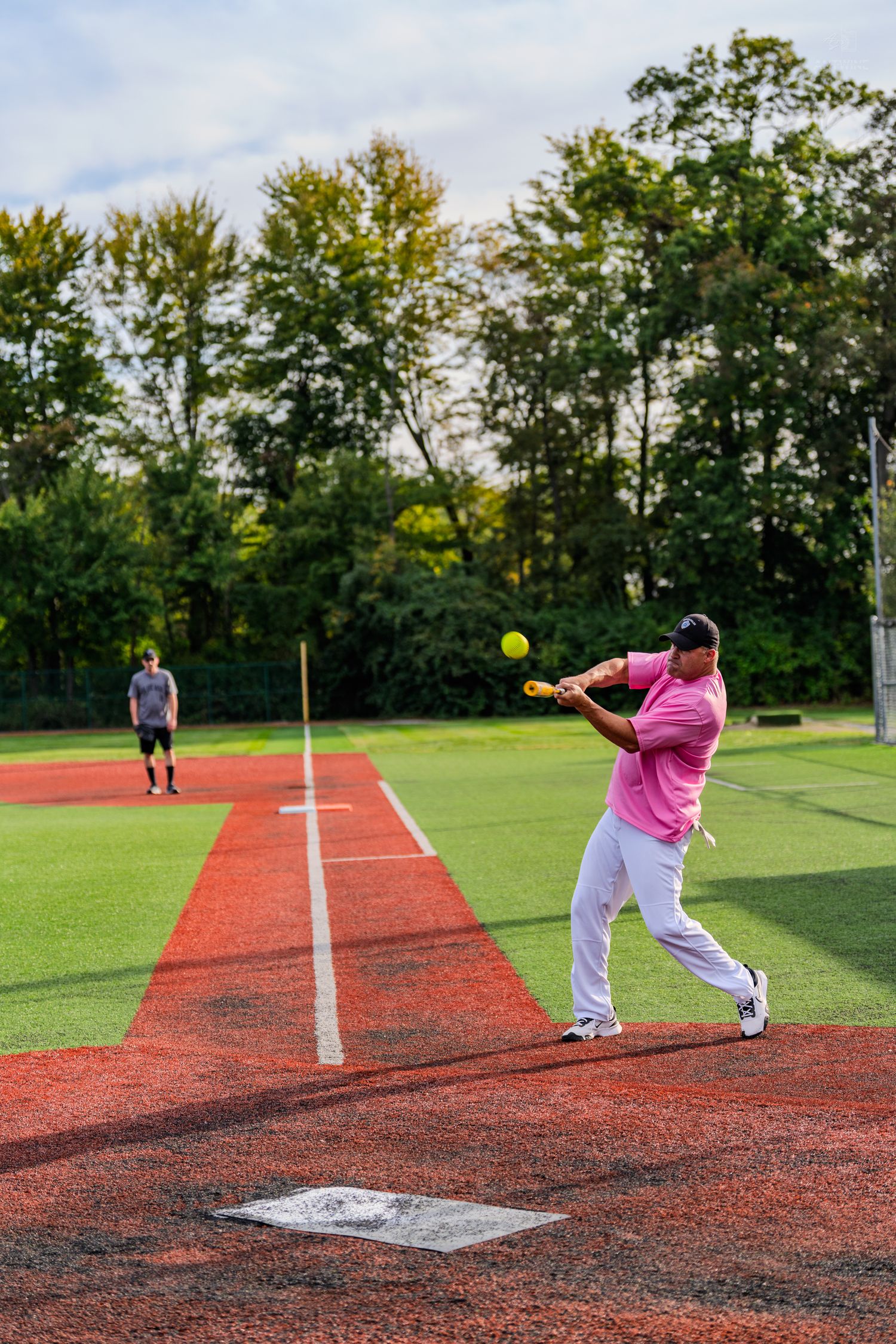 Player in pink shirt hitting yellow softball on red artificial turf field during outdoor game.