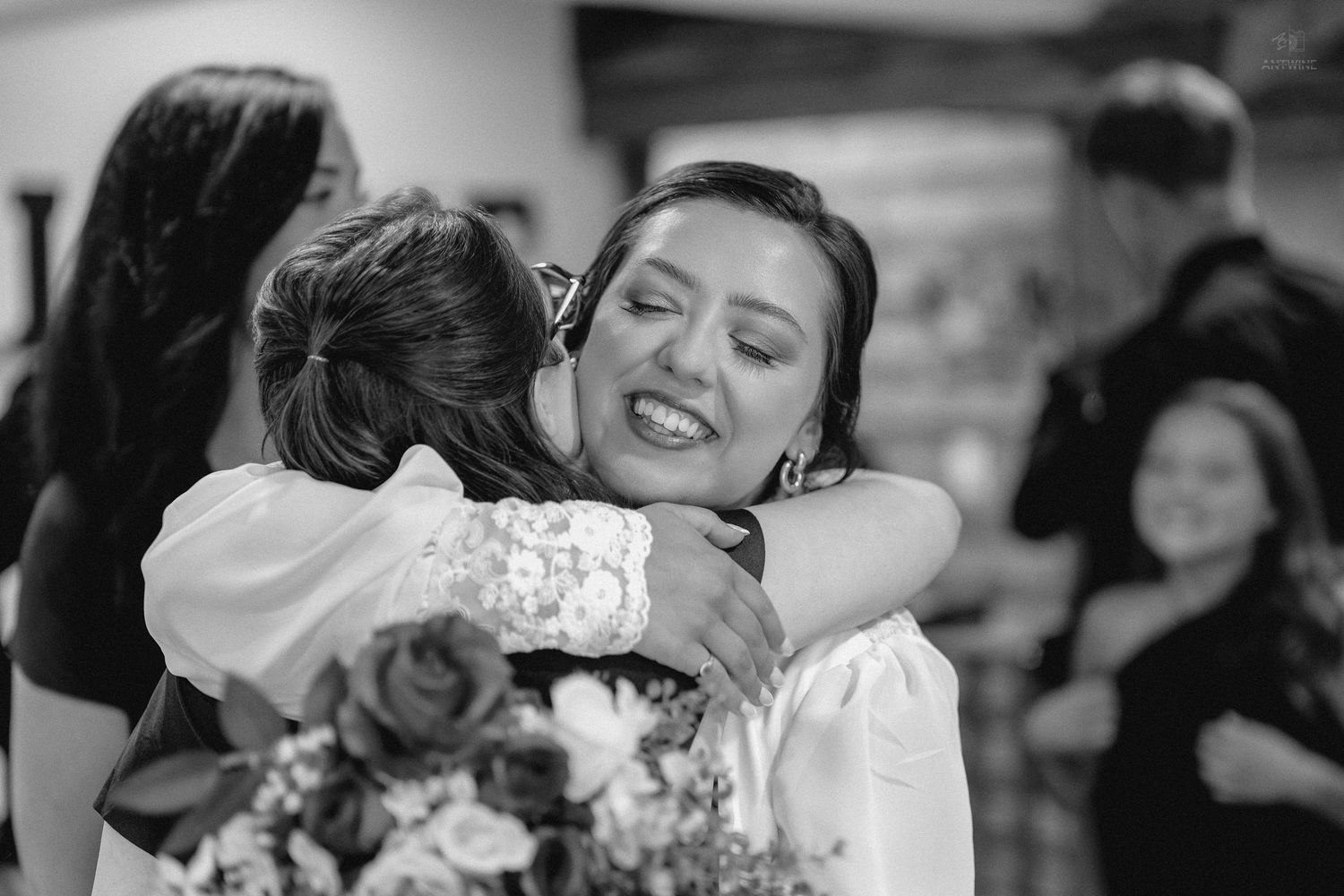 Bride in white lace wedding dress joyfully embraces a guest while holding a floral bouquet.
