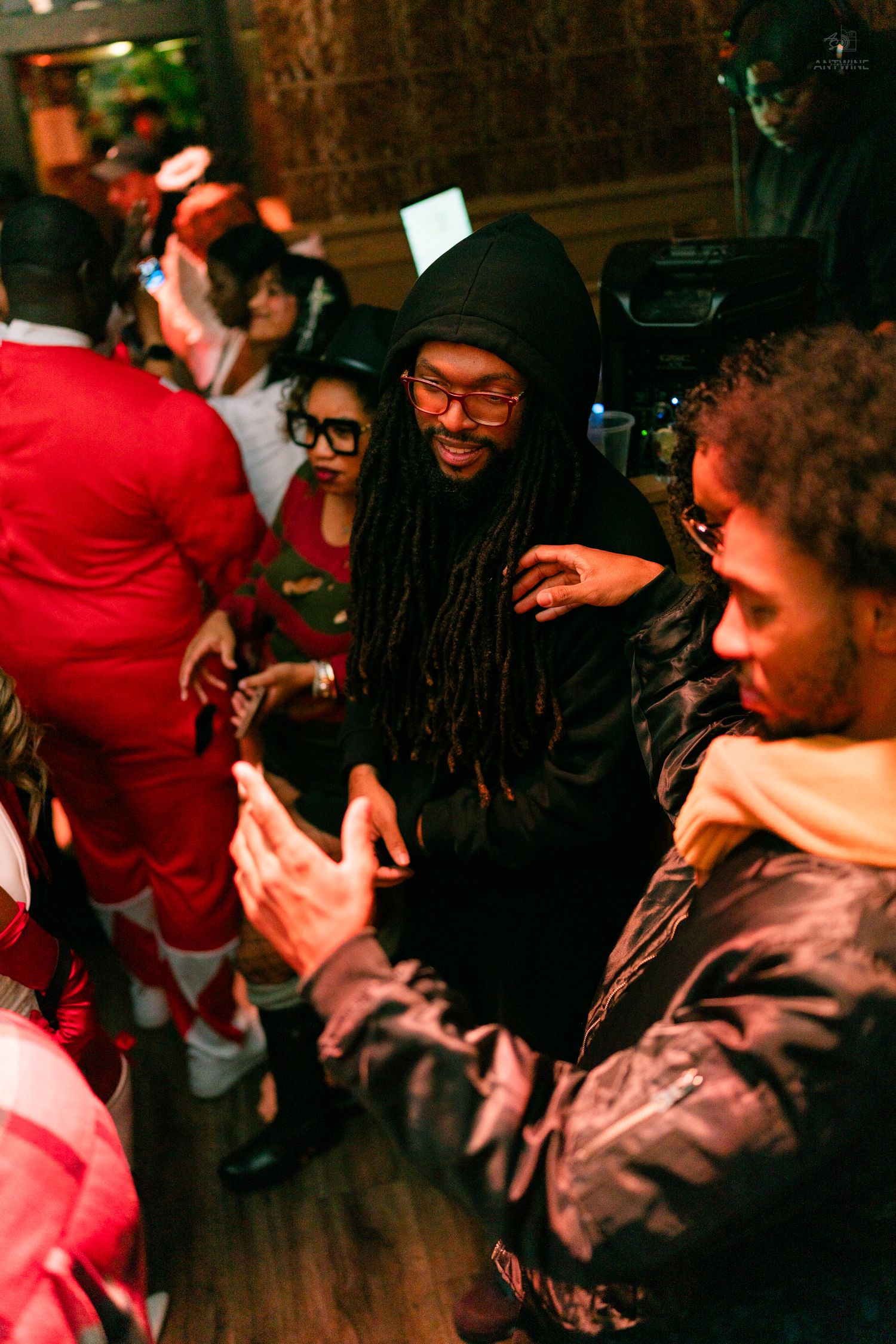 Man with dreadlocks in a black hoodie smiling and dancing at a Halloween costume party indoors.