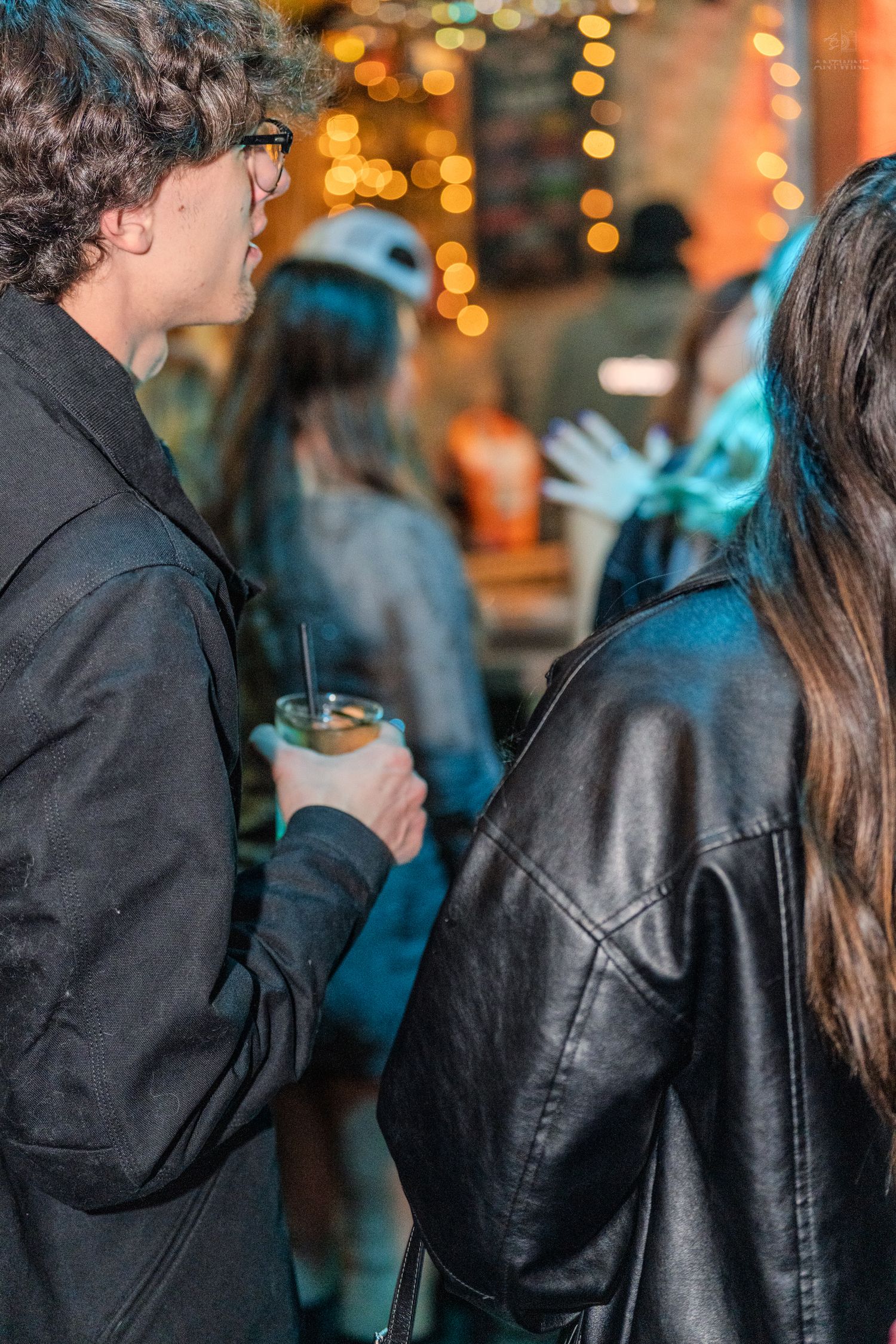 Two young people in black jackets socializing at a lively bar with warm bokeh lights in the background.