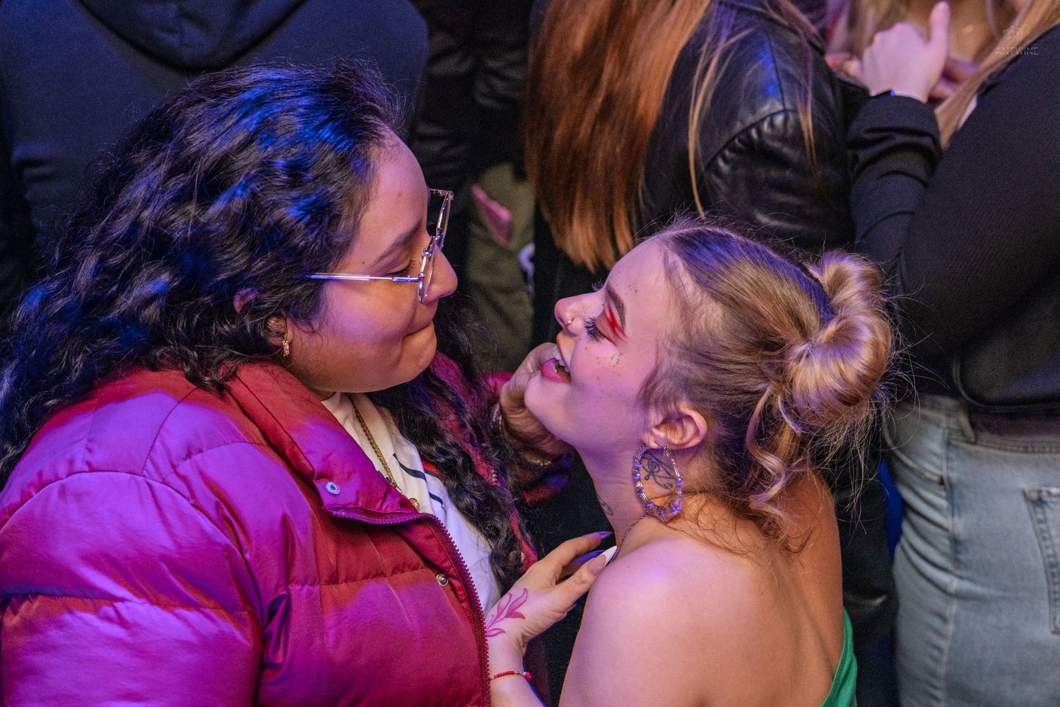 Two women interact closely at a nightclub event under blue and purple lighting.