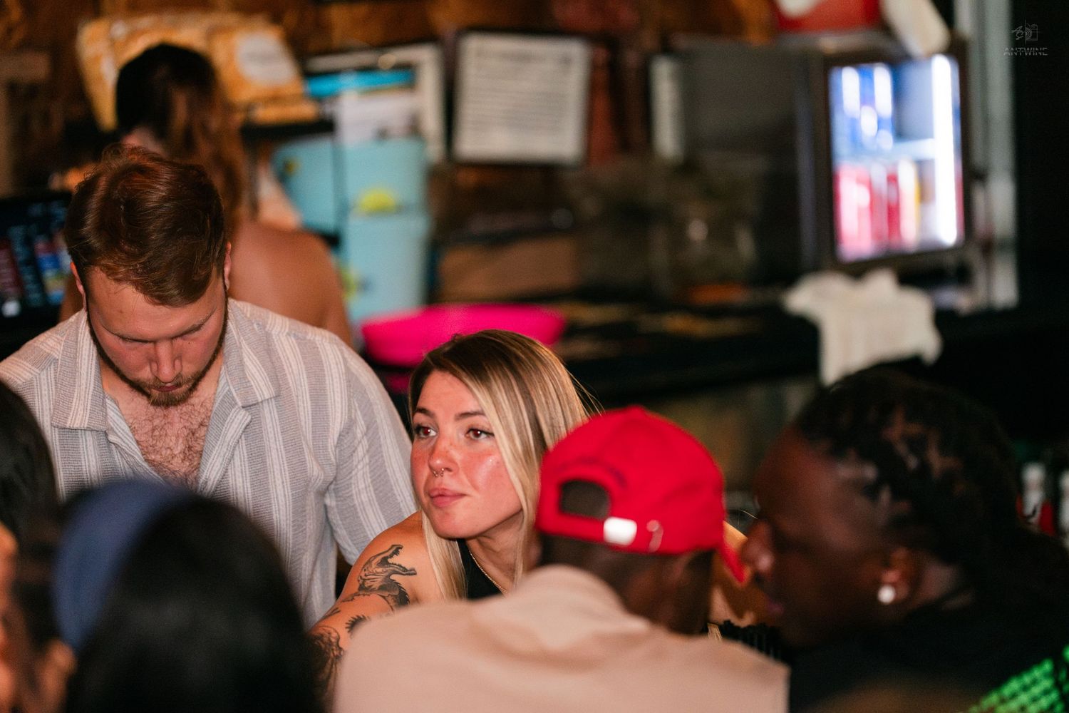 Crowded bar scene with a blonde woman and tall man in a striped shirt socializing indoors.