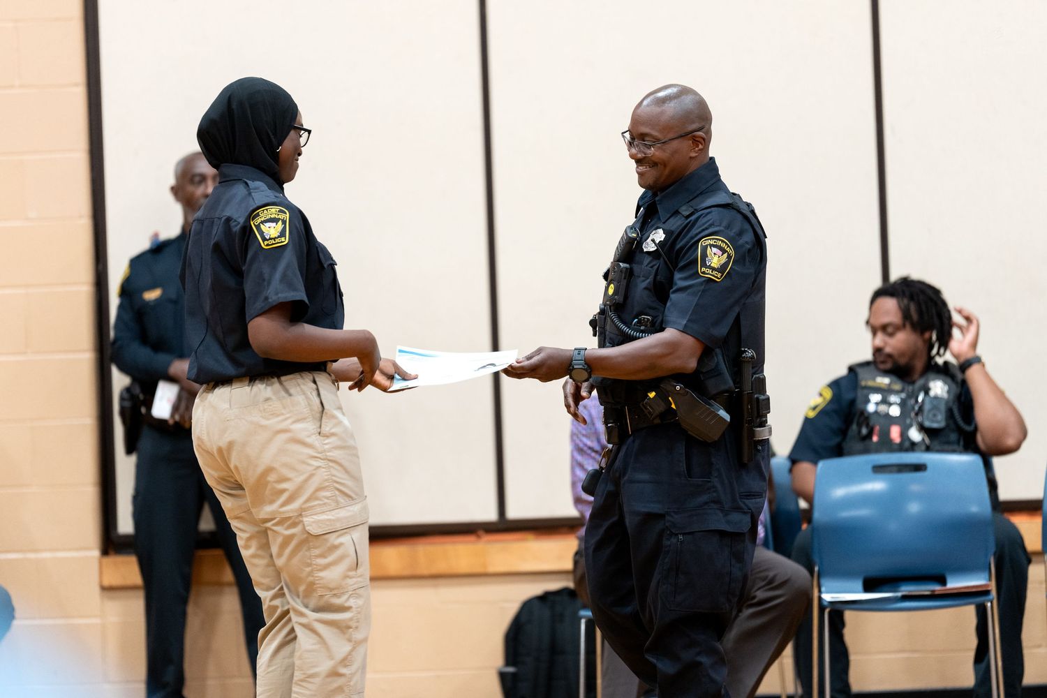 Two police officers in uniform exchange documents at an indoor community event with others visible in the background.