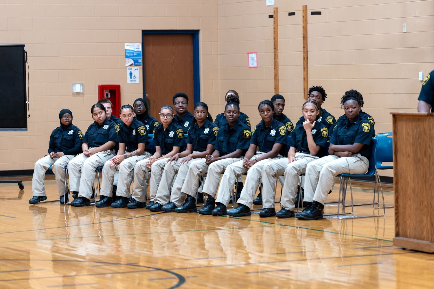 Students in uniform shirts and khaki pants sit in chairs during a school assembly in a gymnasium.
