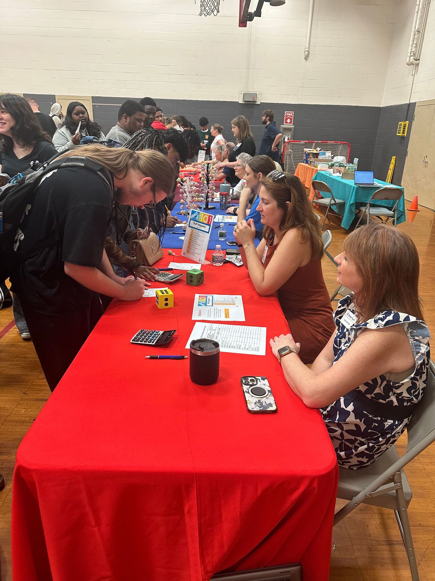 Students and recruiters interact at a school career fair in a gymnasium with colorful tables.