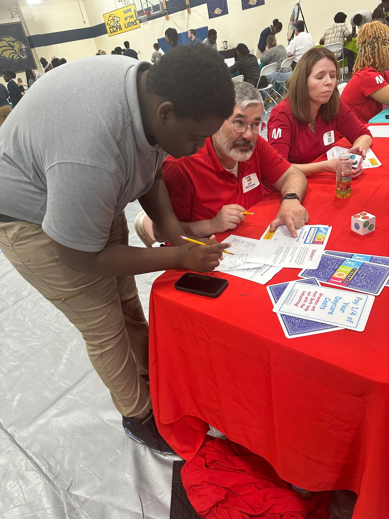 Student leans over red table signing papers with two volunteers in red shirts at an educational event.