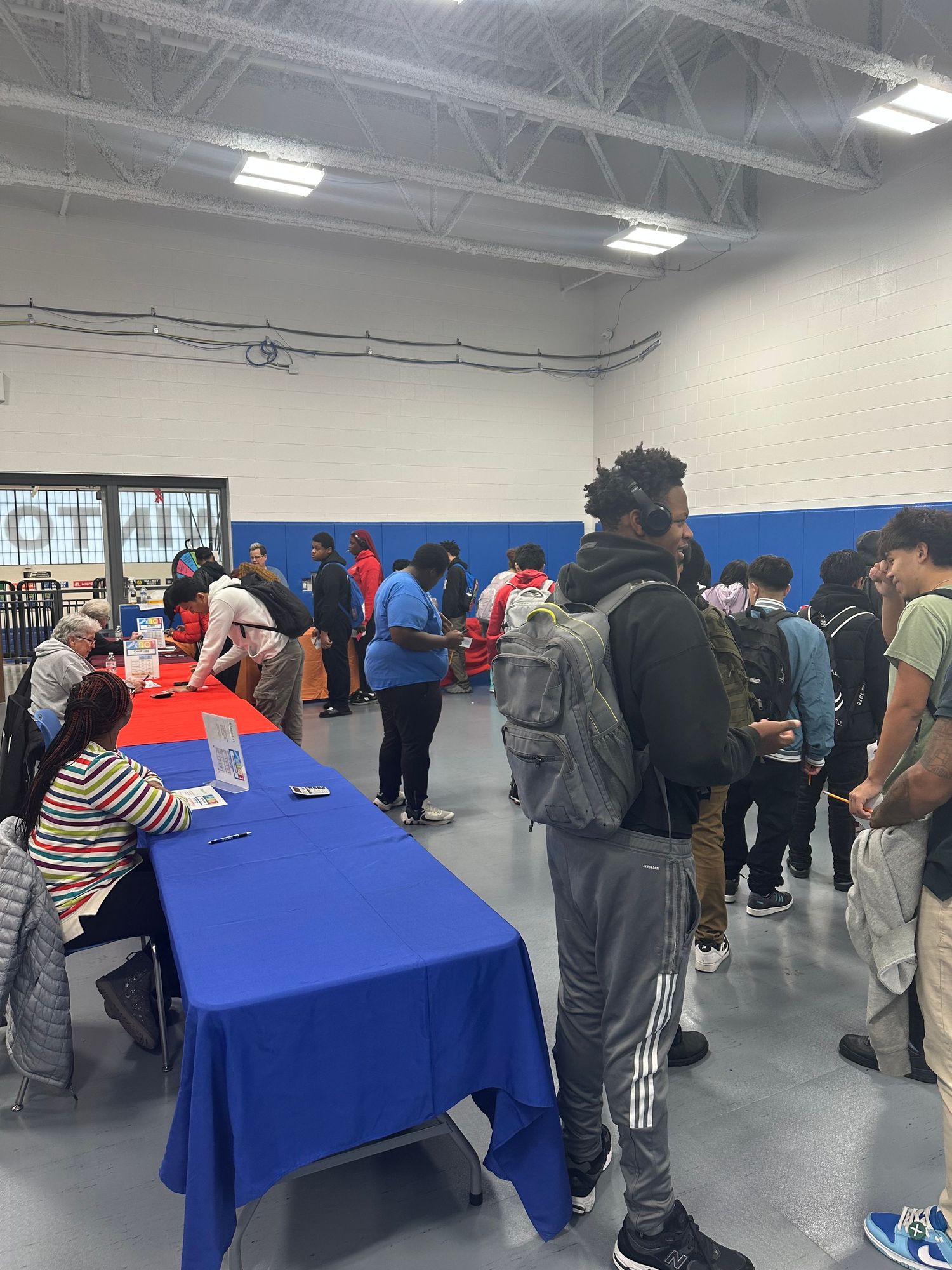 Diverse students line up at blue-clothed tables during a school career fair event held in a gymnasium.