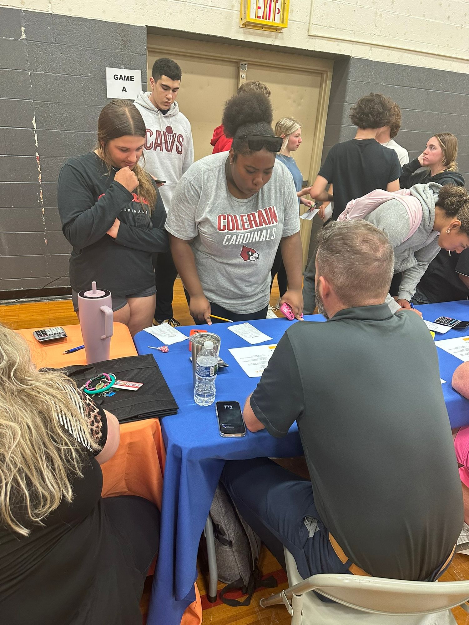 High school students participate in a financial literacy activity at a blue table in a gymnasium.
