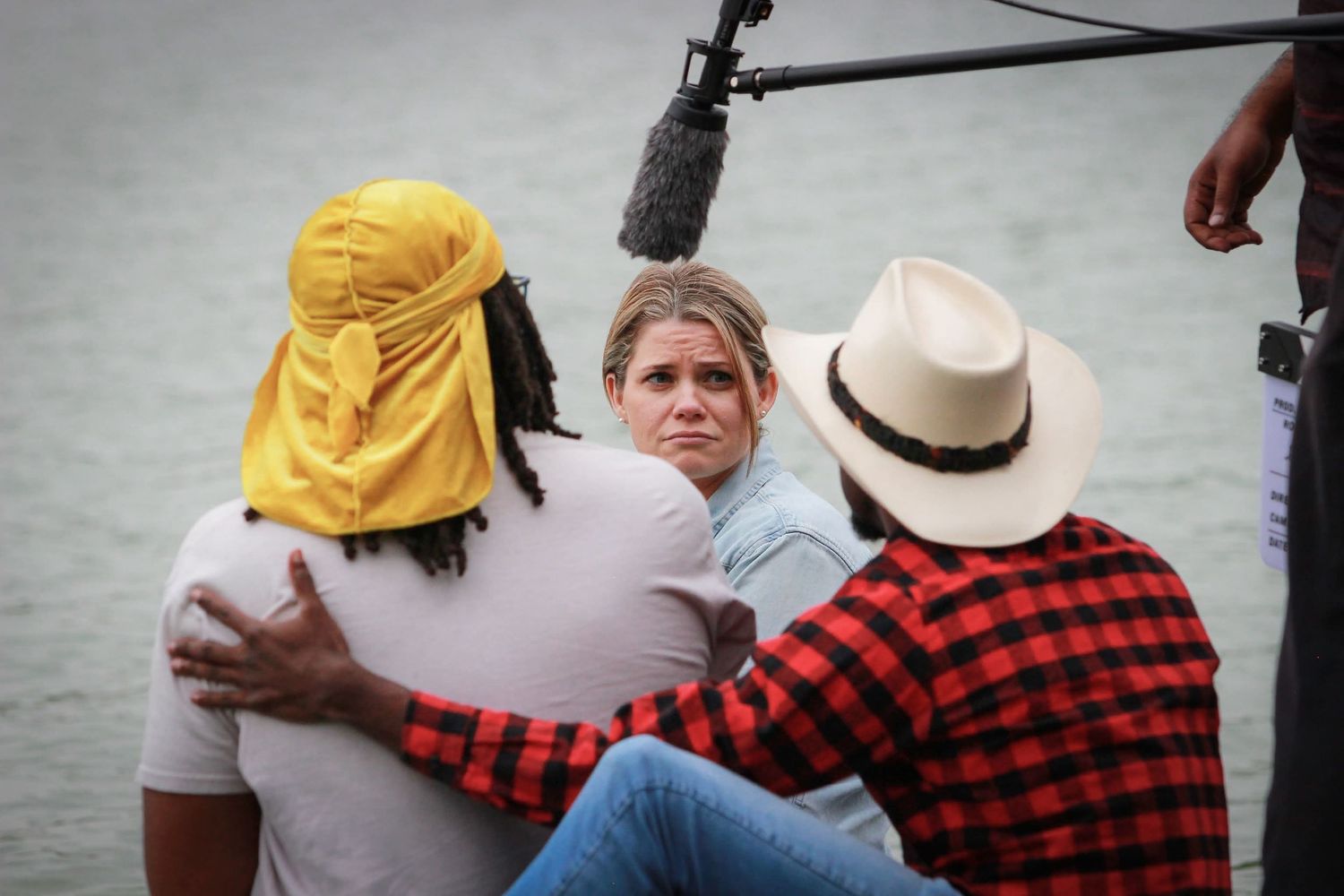Three people sit together near water on a film set with a boom microphone overhead in an outdoor scene.
