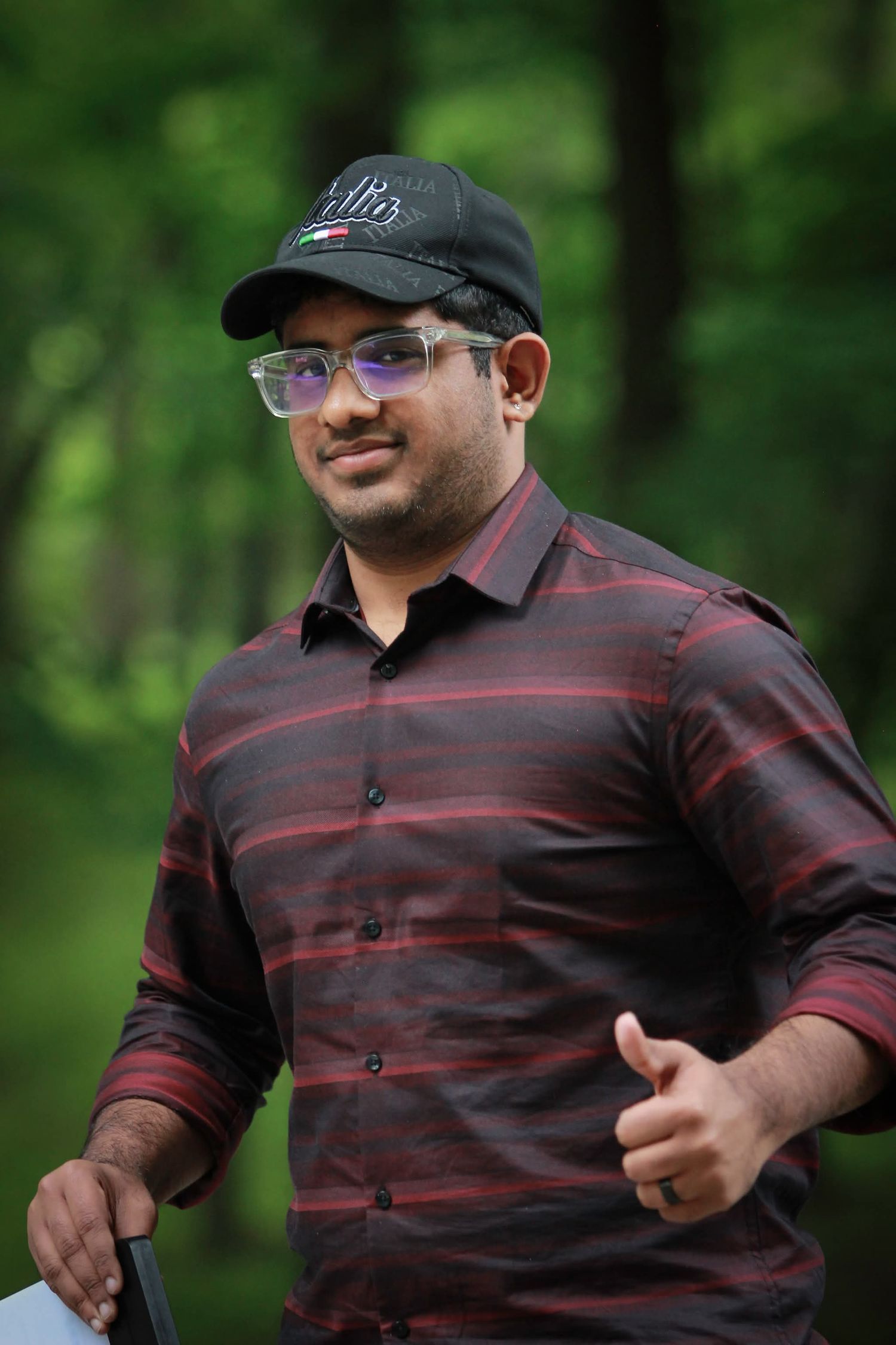 Smiling man wearing glasses and black cap holding a tablet outdoors in a green forest.
