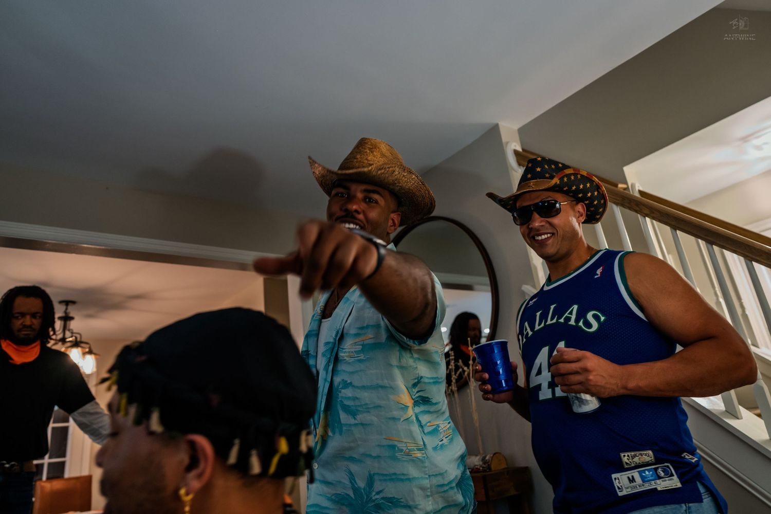 Two men in cowboy hats smiling and pointing at a casual indoor house party.