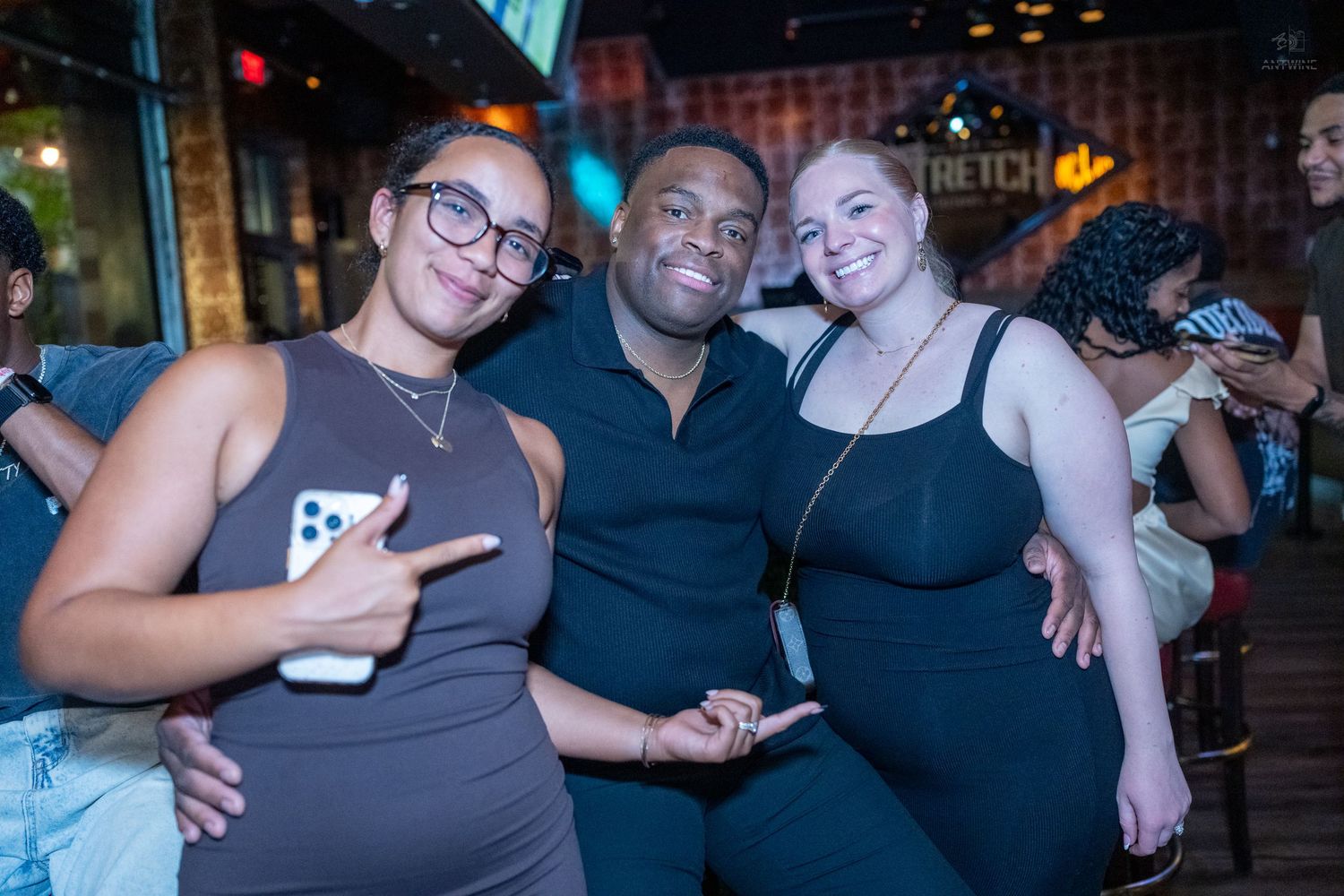 Three friends smile and pose together at a bar with warm ambient lighting behind them.