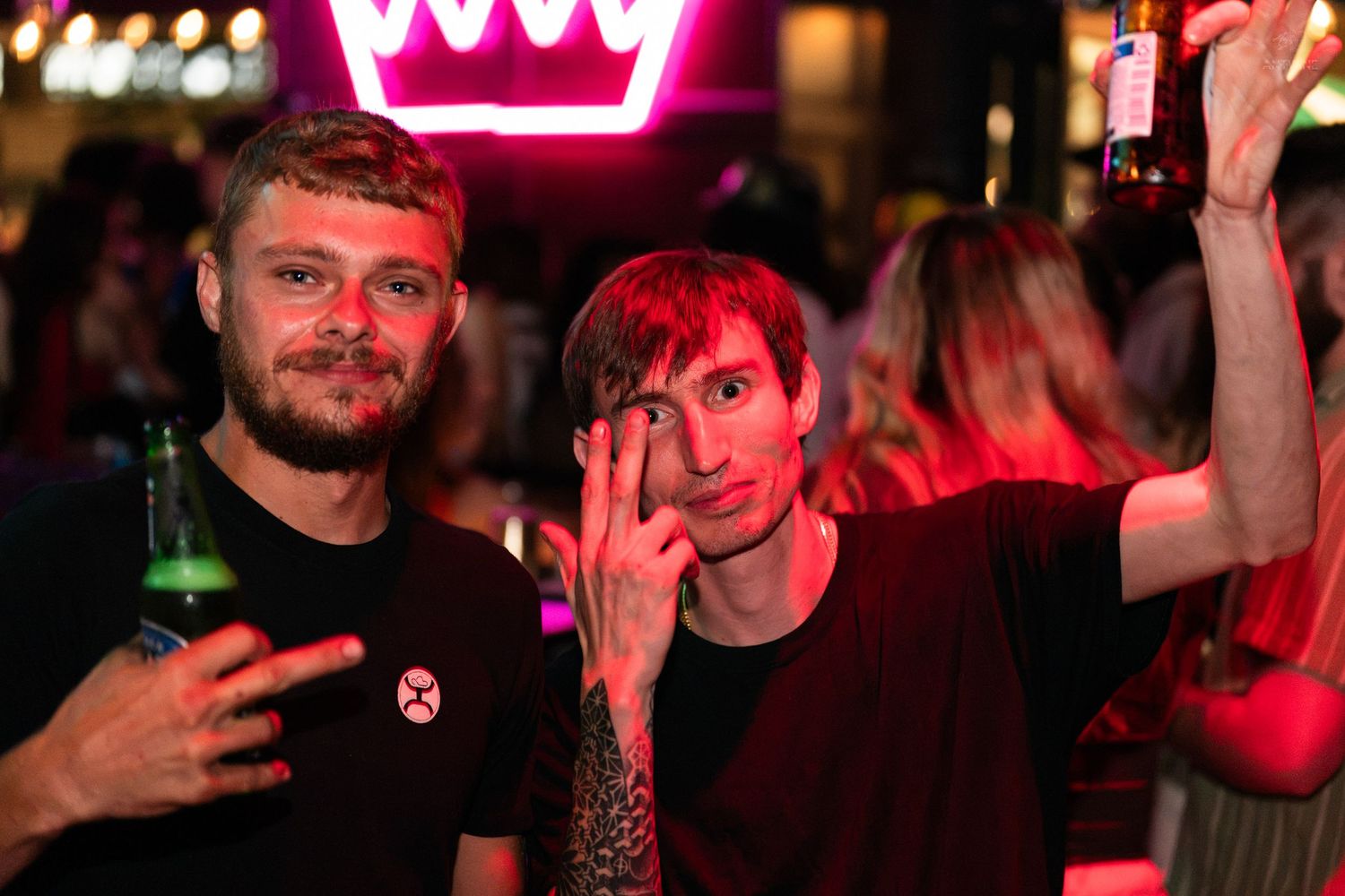 Two men in black shirts making hand gestures and smiling under a crown-shaped neon sign at a bar.