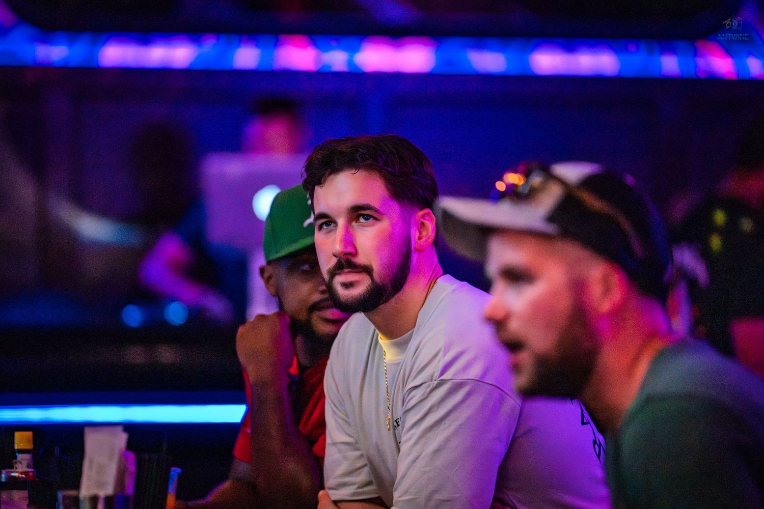 Two men watch intently under colorful neon lights at a bar or nightclub.