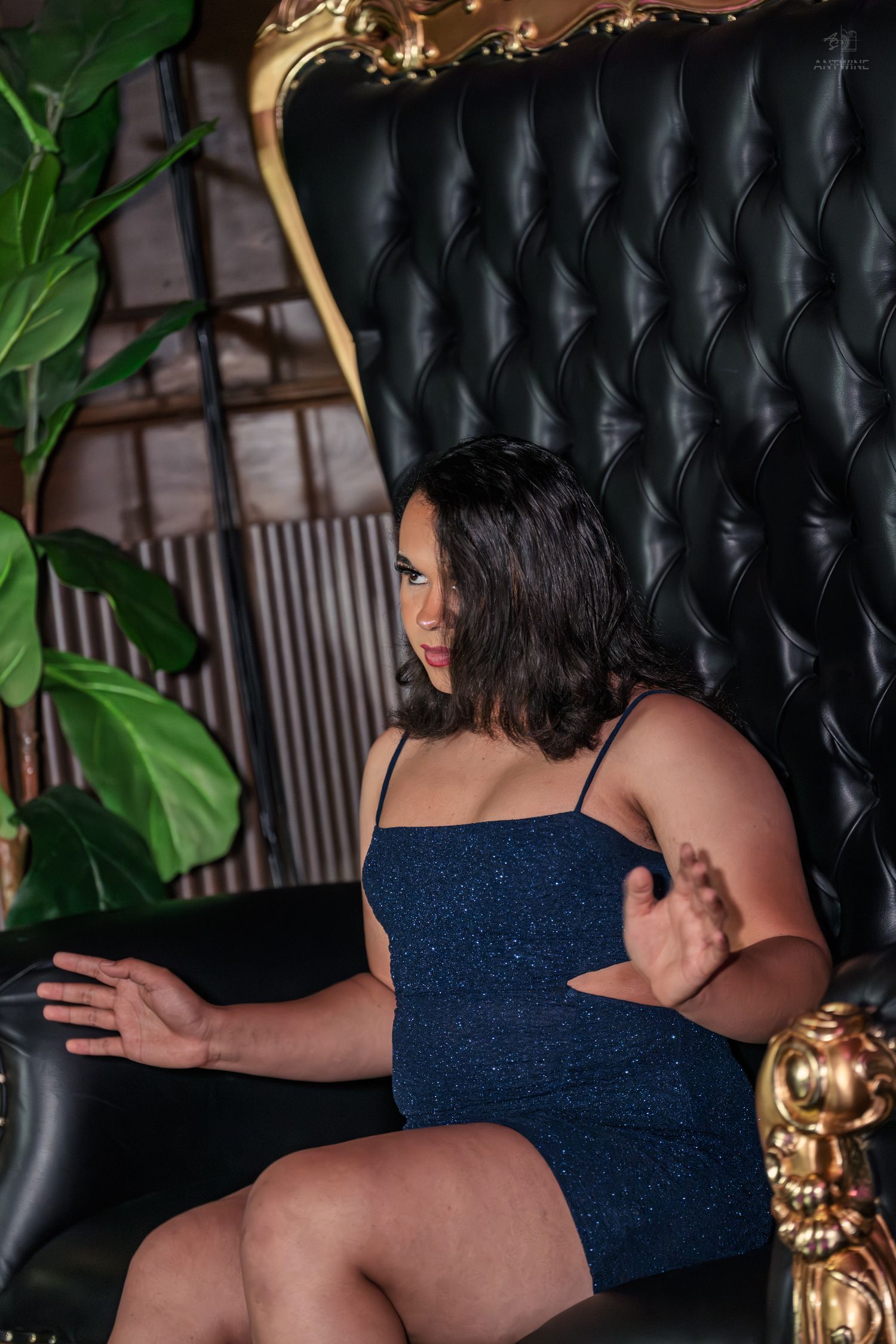 Woman in navy blue sparkle mini dress poses on ornate black tufted throne chair with gold accents.
