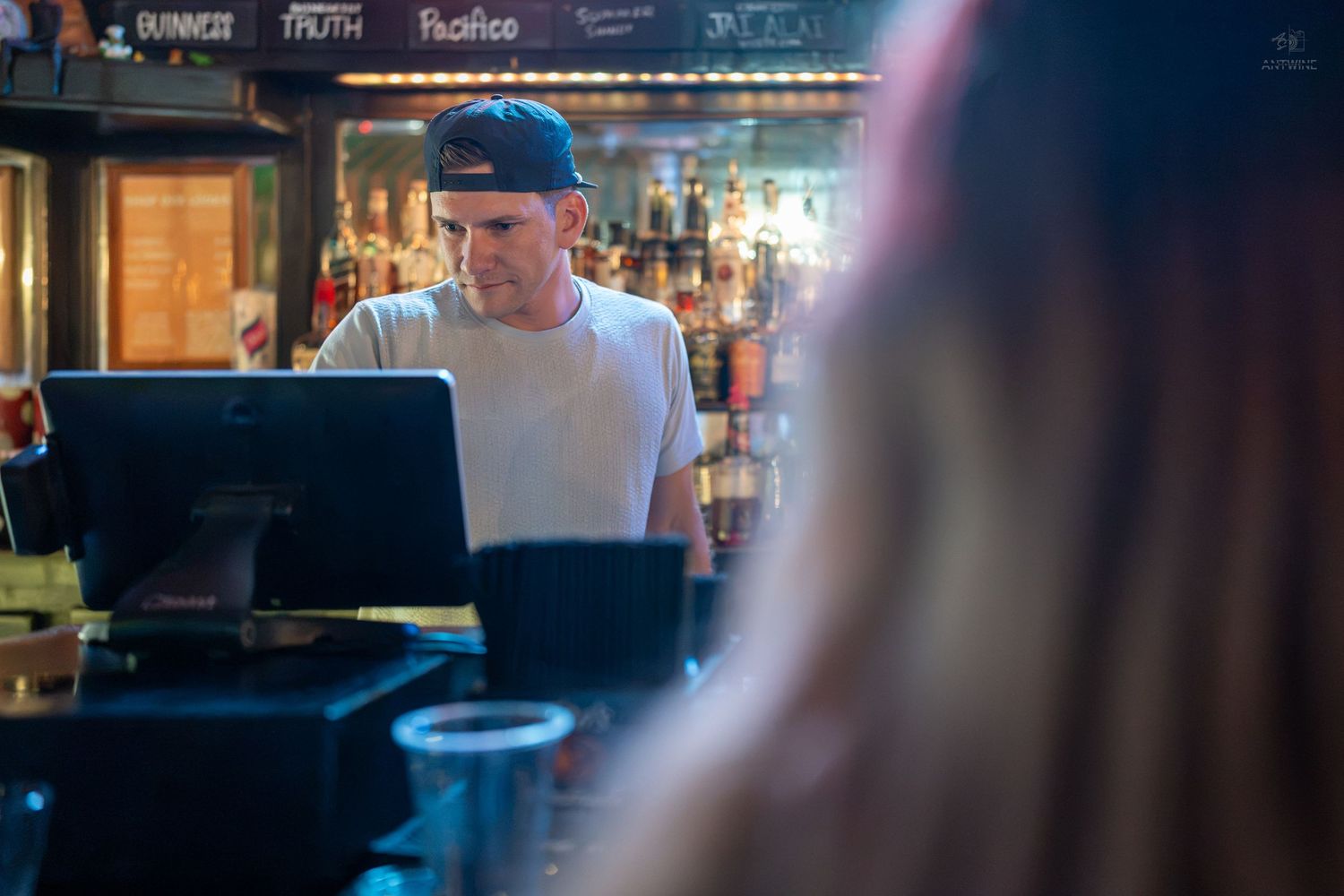 Bartender in a cap works at a POS terminal inside a dimly lit bar with liquor bottles in the background.