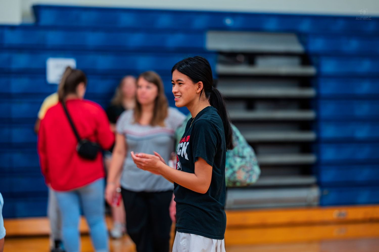 A young woman in a black t-shirt smiles while holding a paper in a school gymnasium.