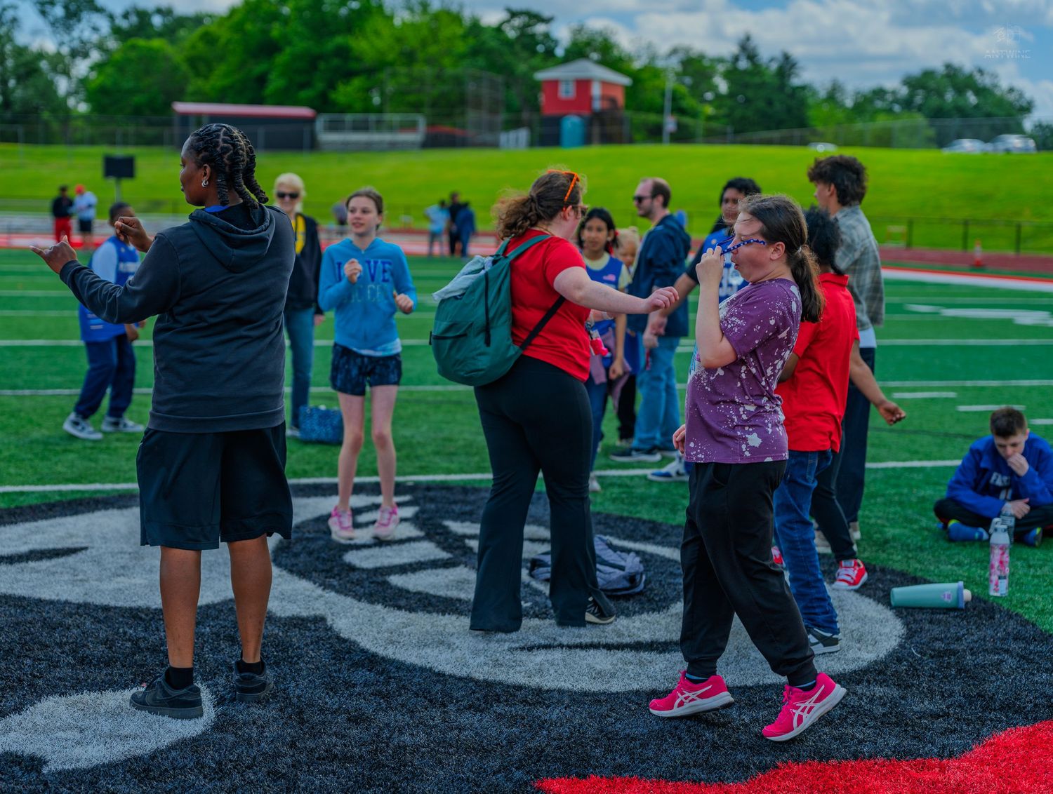 Students and adults gather on a school athletic track field during an outdoor event on a cloudy day.