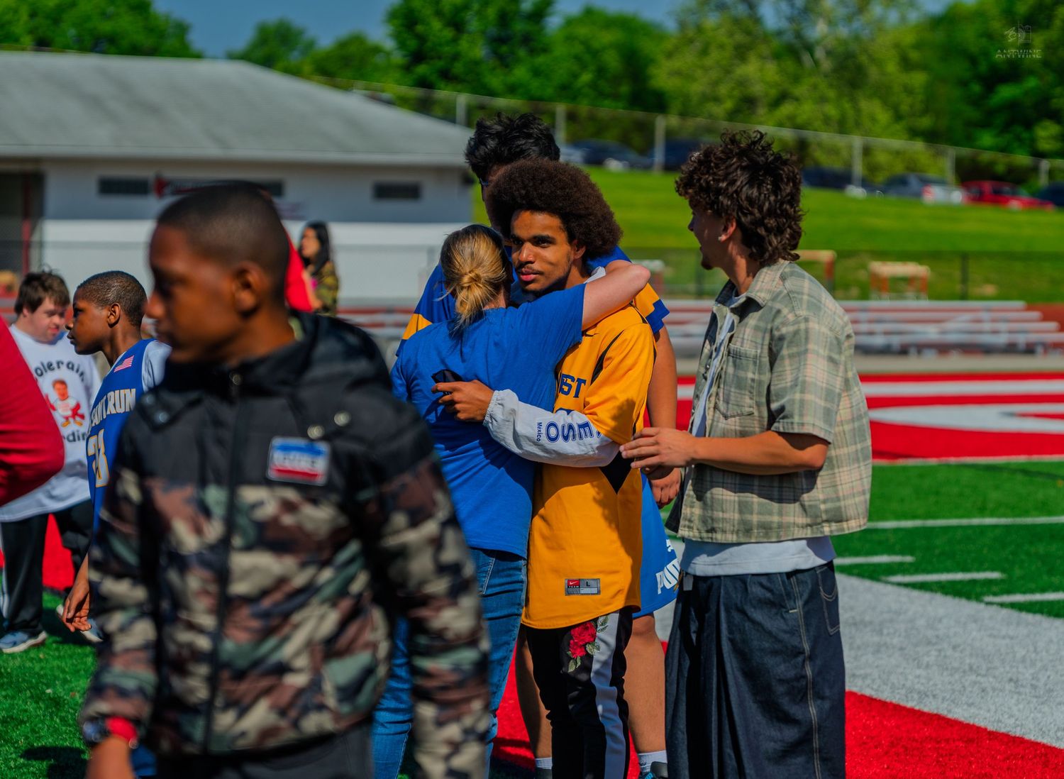 Young students hugging and socializing on a red track field during an outdoor school event on a sunny day.