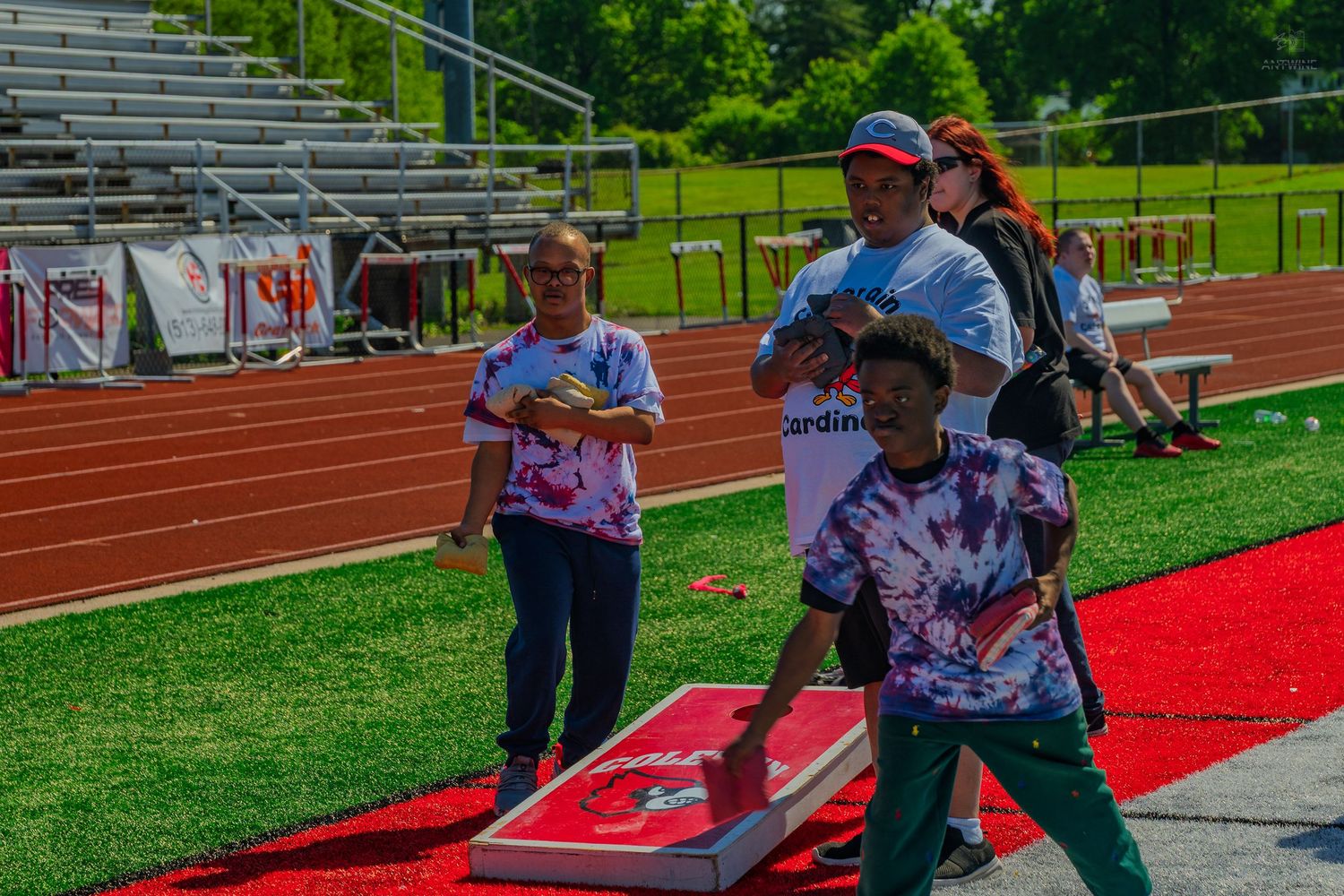 People playing cornhole on a red track field during an outdoor school event on a sunny day.