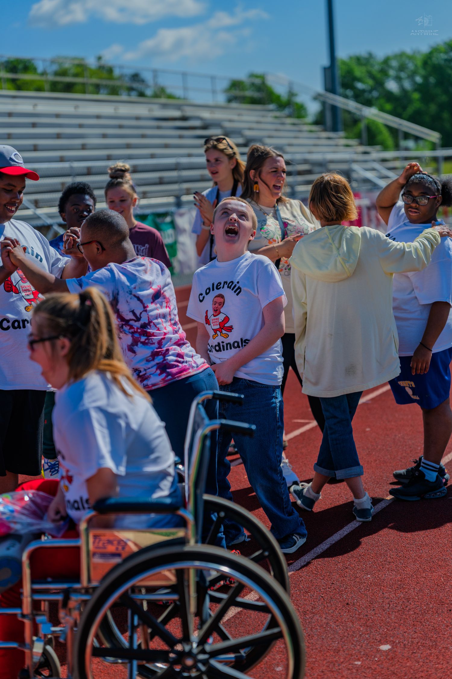 Children and adults celebrate outdoors at a Special Olympics track event, with a wheelchair visible in the foreground.