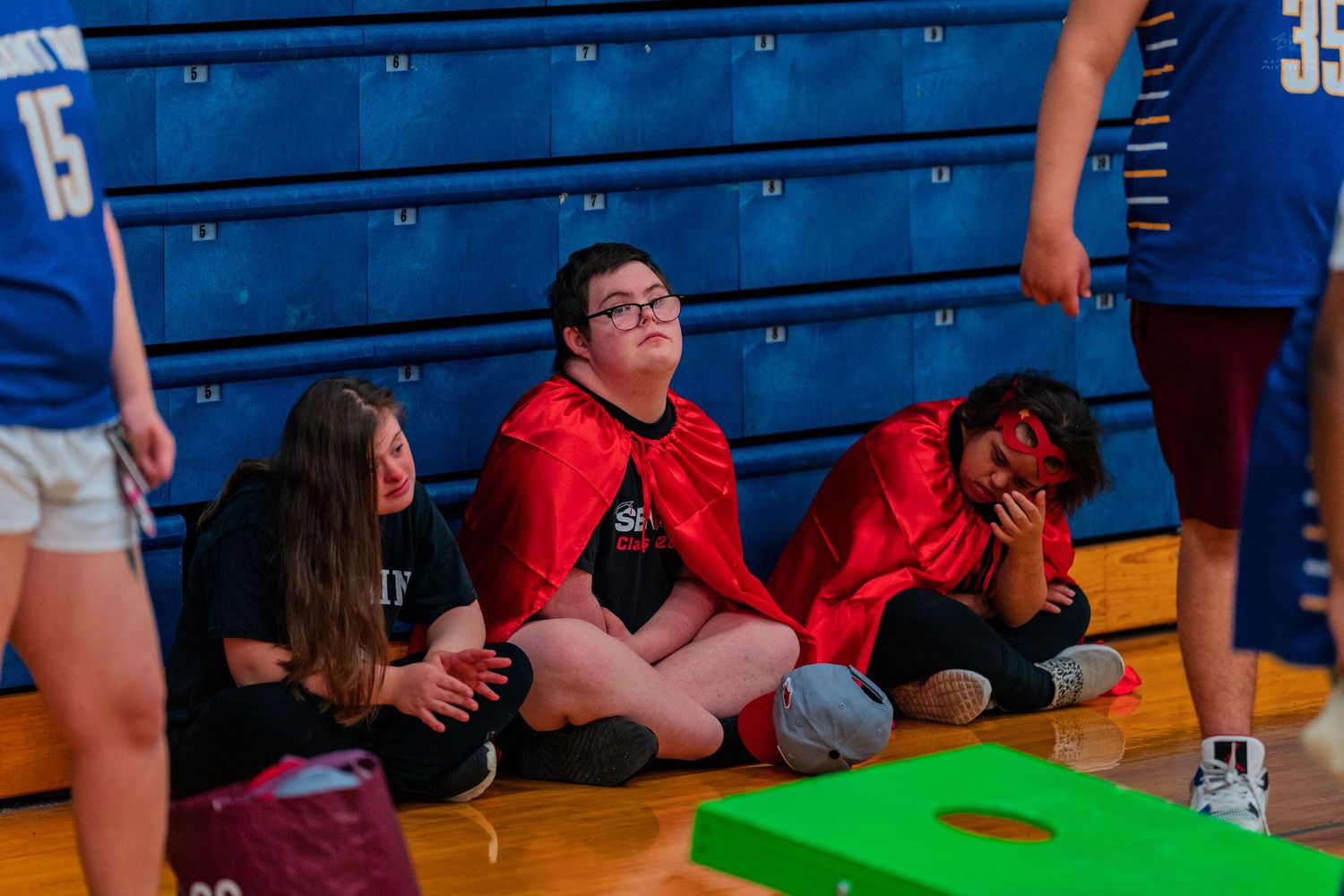 Students in costumes sit on a gym floor during a school event, one wearing a red cape, with cornhole boards nearby.