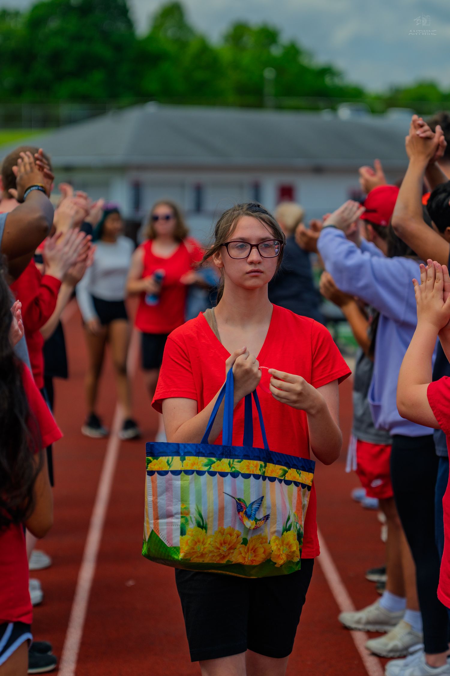Young girl in red shirt walks through cheering crowd on track, holding colorful tote bag at school sports event.