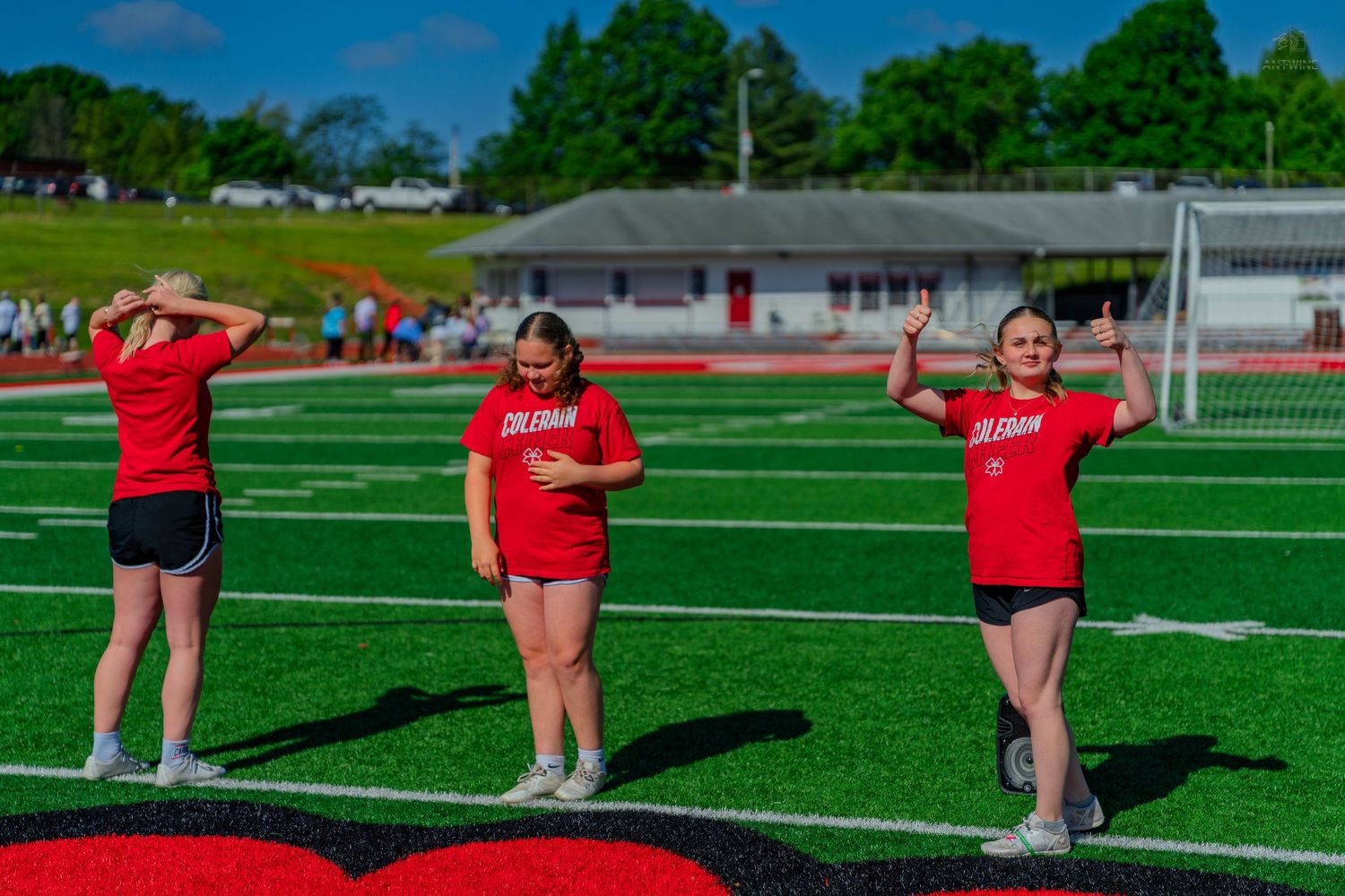Three girls in red Coleman shirts perform cheerleading routines on a green turf football field.