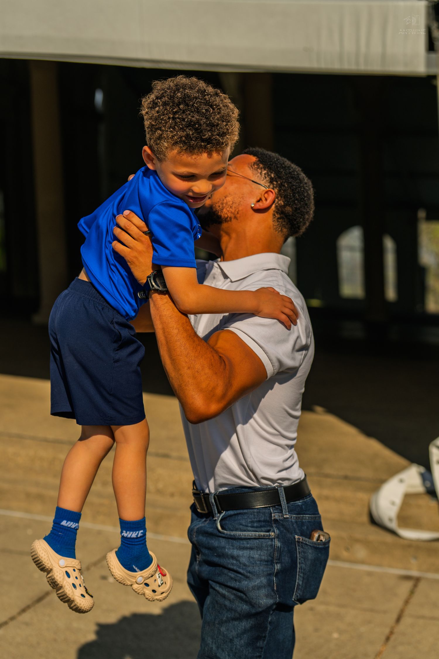 A man lifts a smiling young boy in a blue outfit and kisses him while standing outdoors in sunlight.