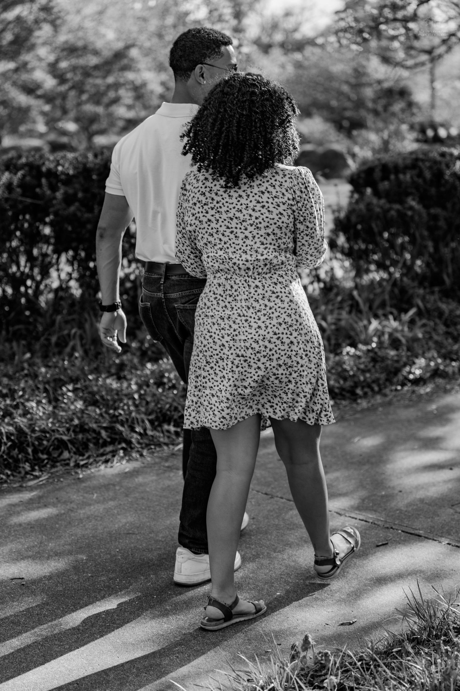 A couple walking closely together on a park path lined with neatly trimmed hedges in black and white.
