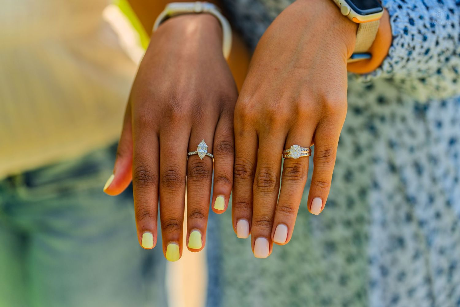 Close-up of two women displaying diamond engagement rings with yellow painted nails outdoors.