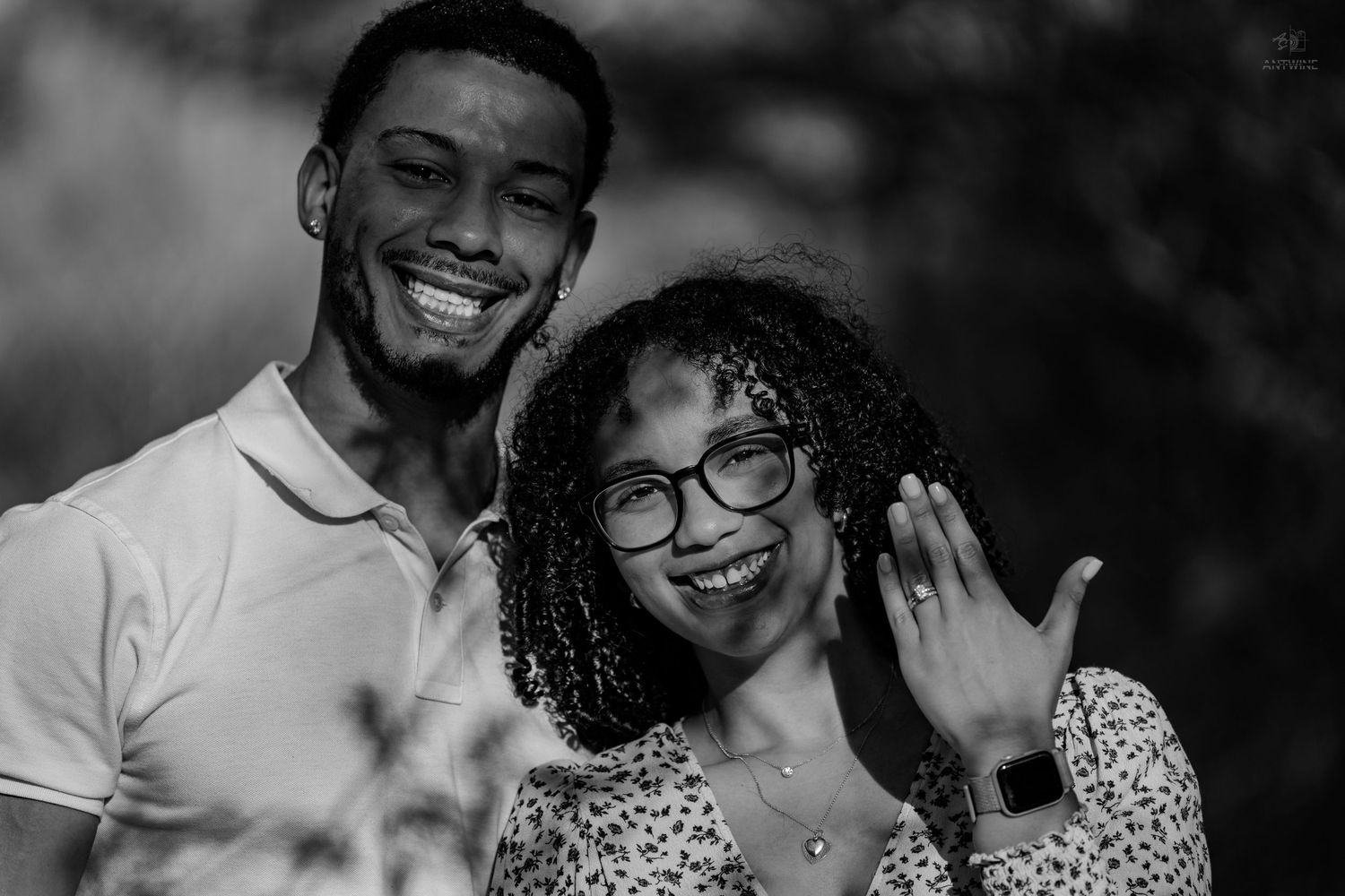 Black and white photo of joyful couple smiling as woman shows off her new engagement ring.