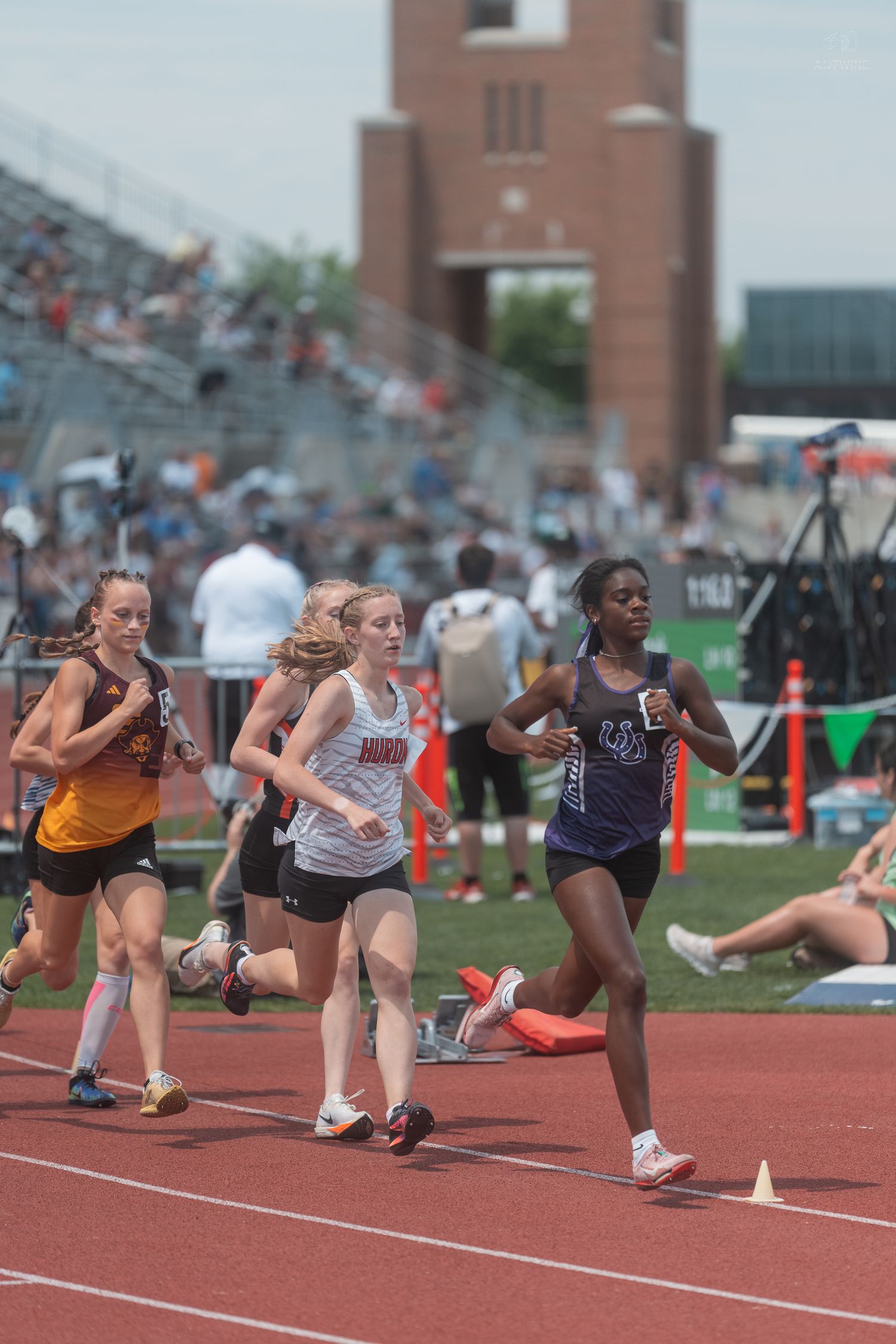 Female high school runners compete on a red track with a brick stadium tower in the background.