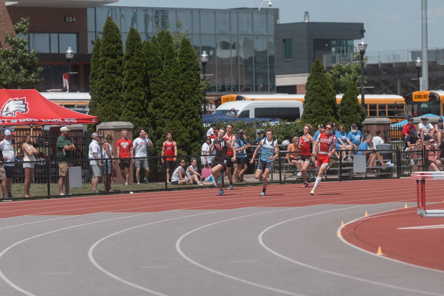 Track athletes rounding a curve during an outdoor high school track meet with spectators lining the field.