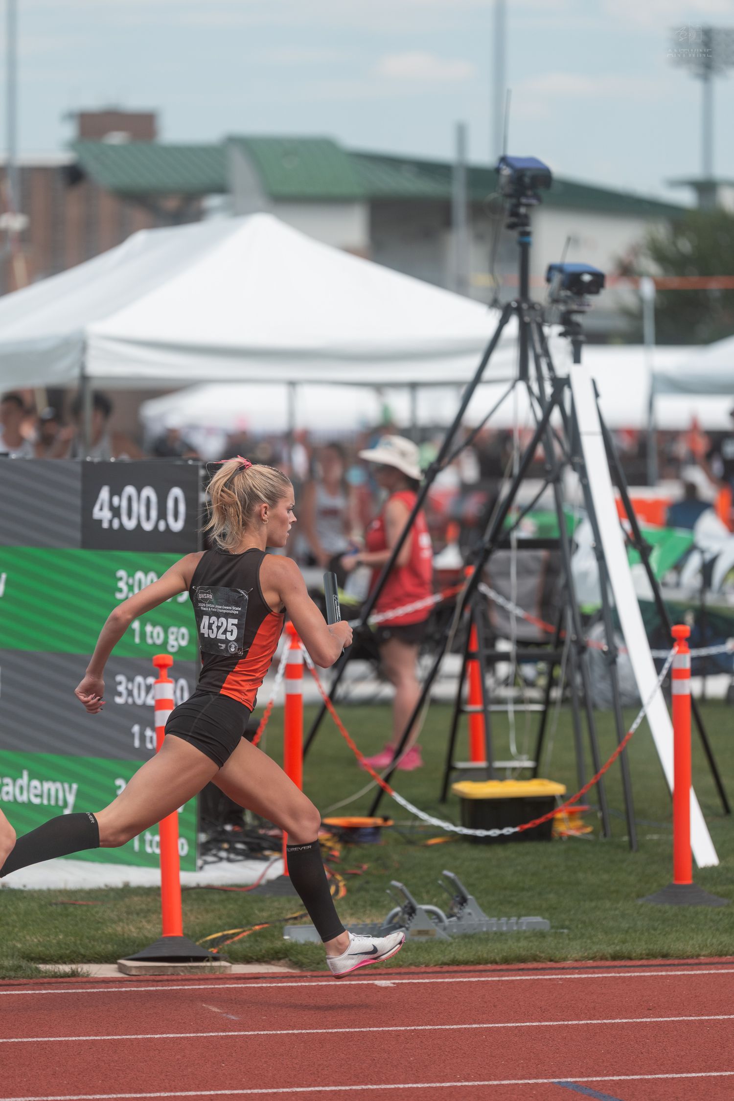 Female track athlete wearing bib 4325 sprints past a timing board showing 4:00.0 during a competitive relay race.
