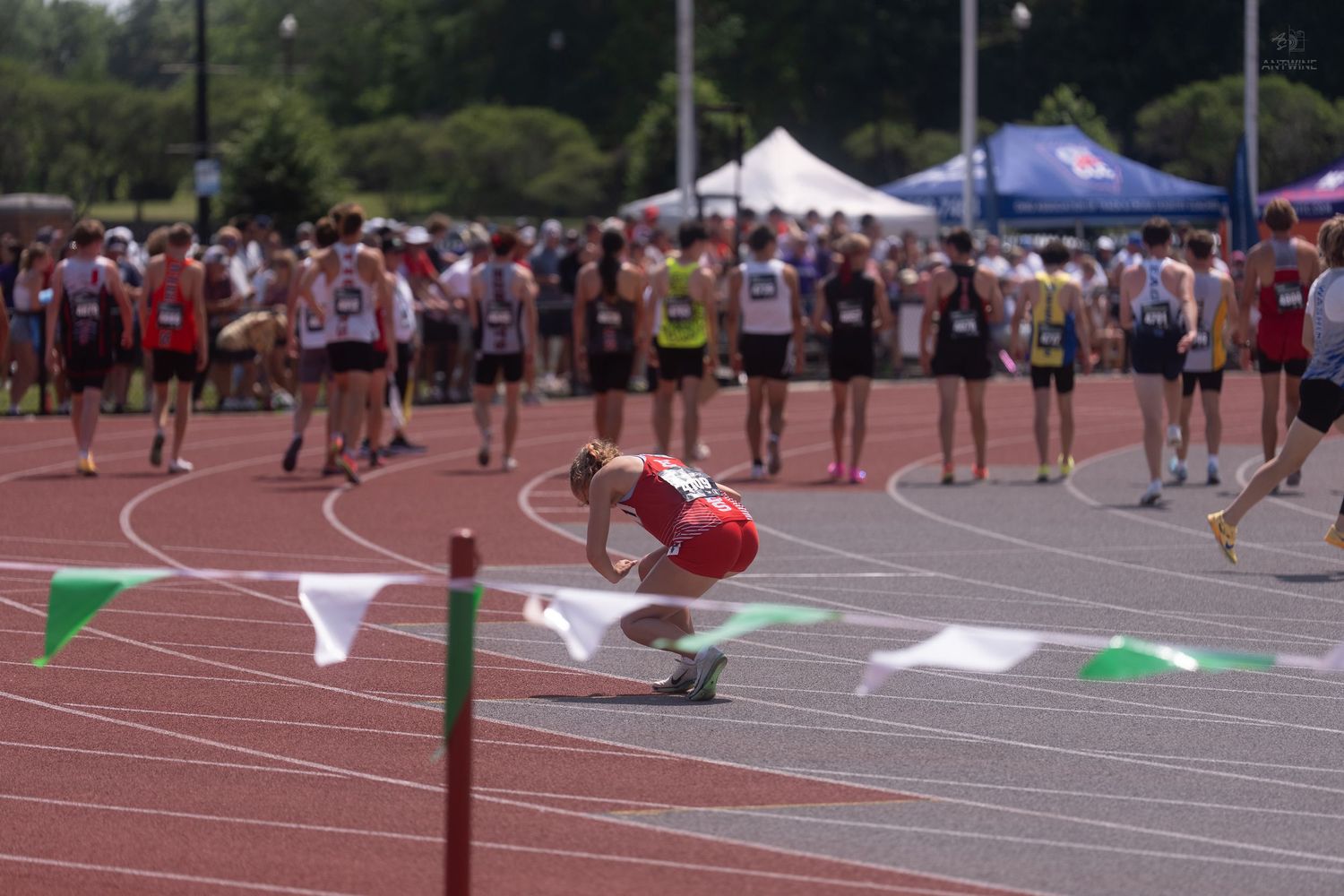 Exhausted runner in red uniform kneels on track after finishing race at crowded meet.