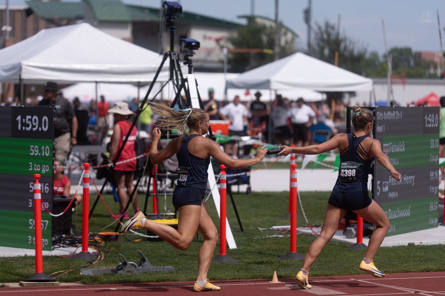 Two female athletes complete a baton exchange during a relay race at a high school state track championship.