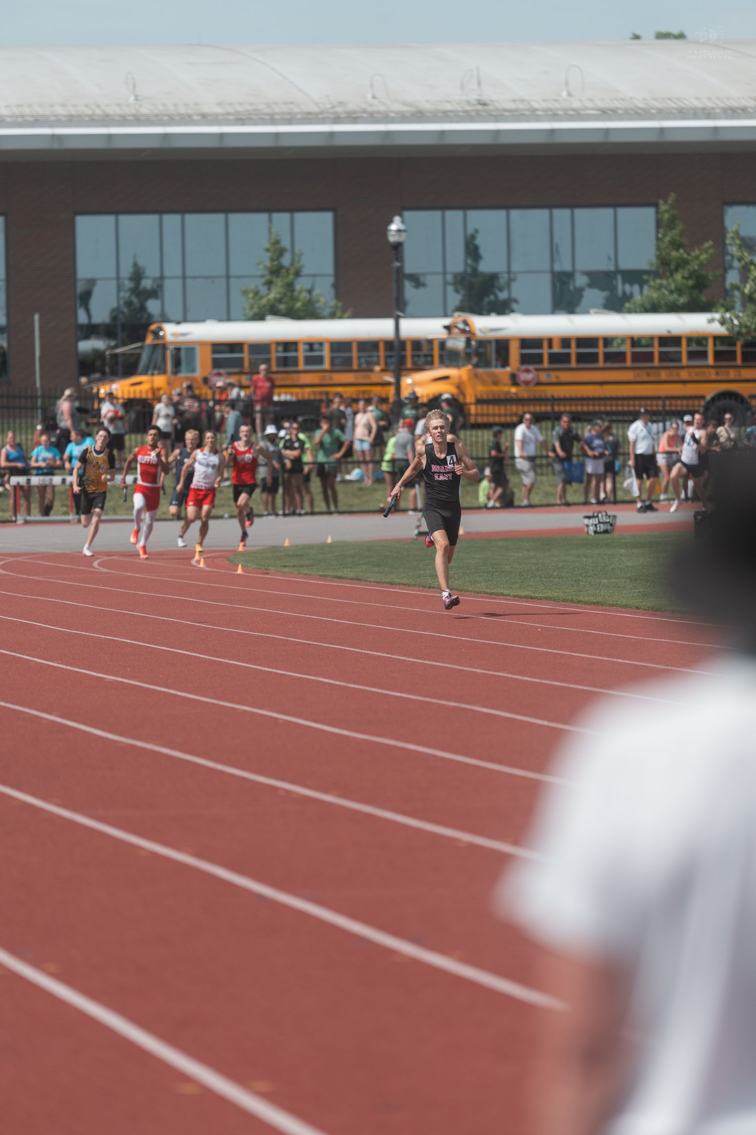 Male runners compete in a track race with yellow school buses and spectators visible in the background.