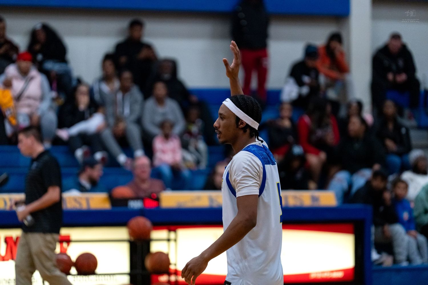 A basketball player in blue and white uniform raises hand signaling during a high school game with packed bleachers behind him.