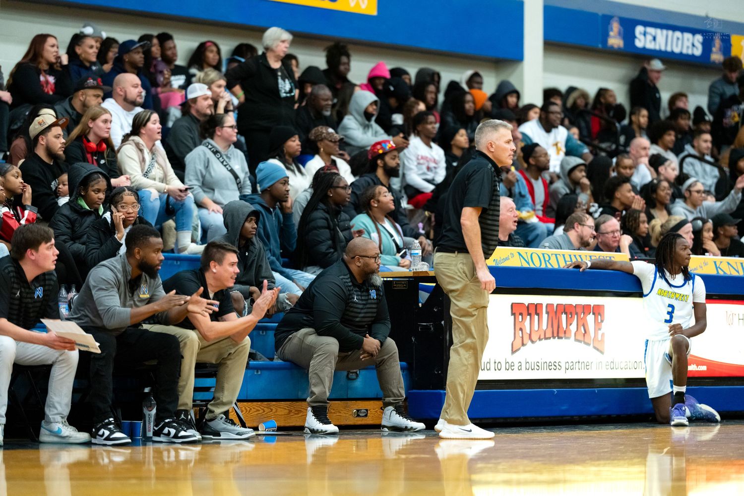 High school basketball coach stands at sideline observing game while team sits on bench behind him.