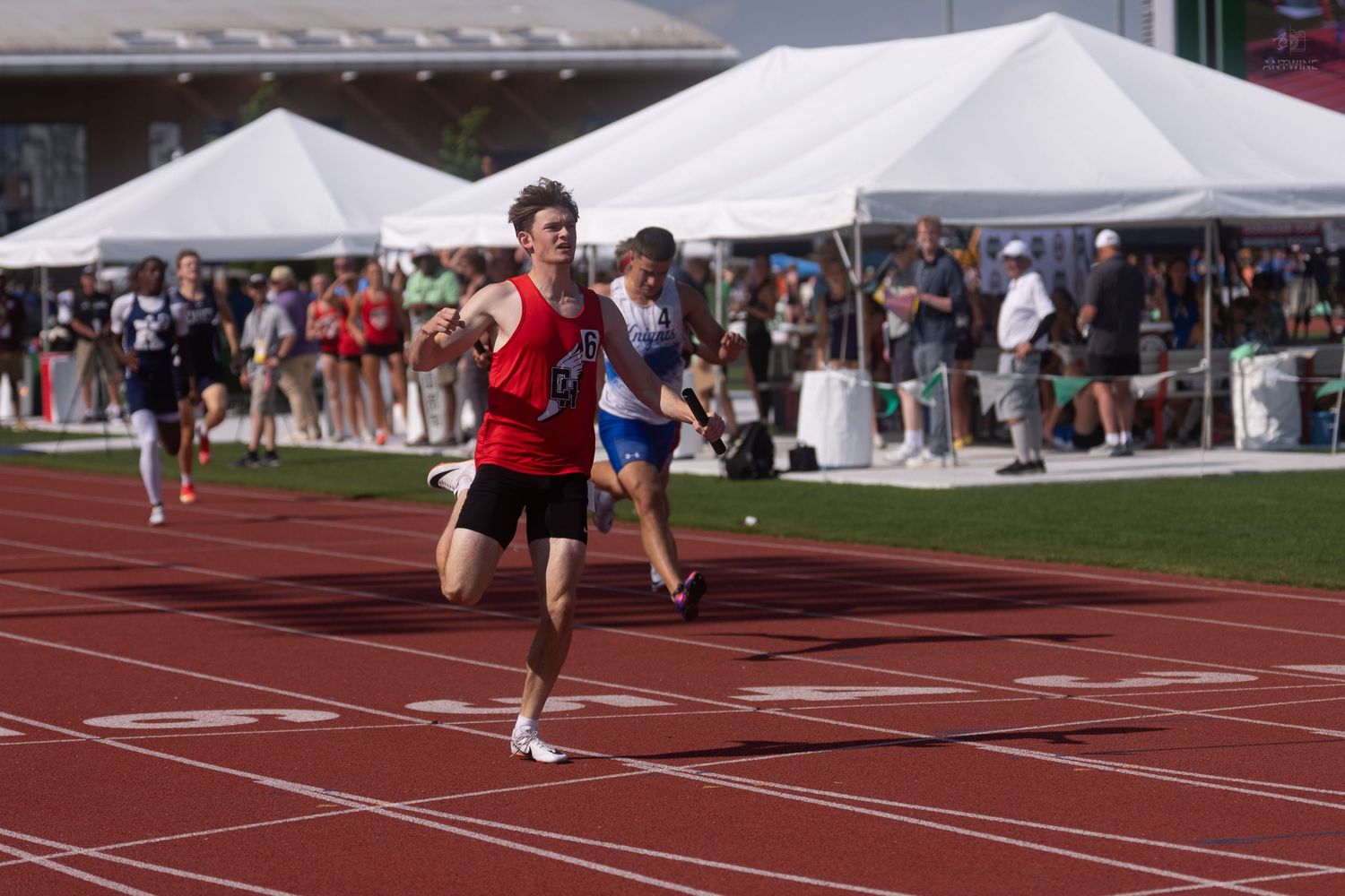 Athlete in red uniform pulls ahead of competitors near the finish line at an outdoor track meet.