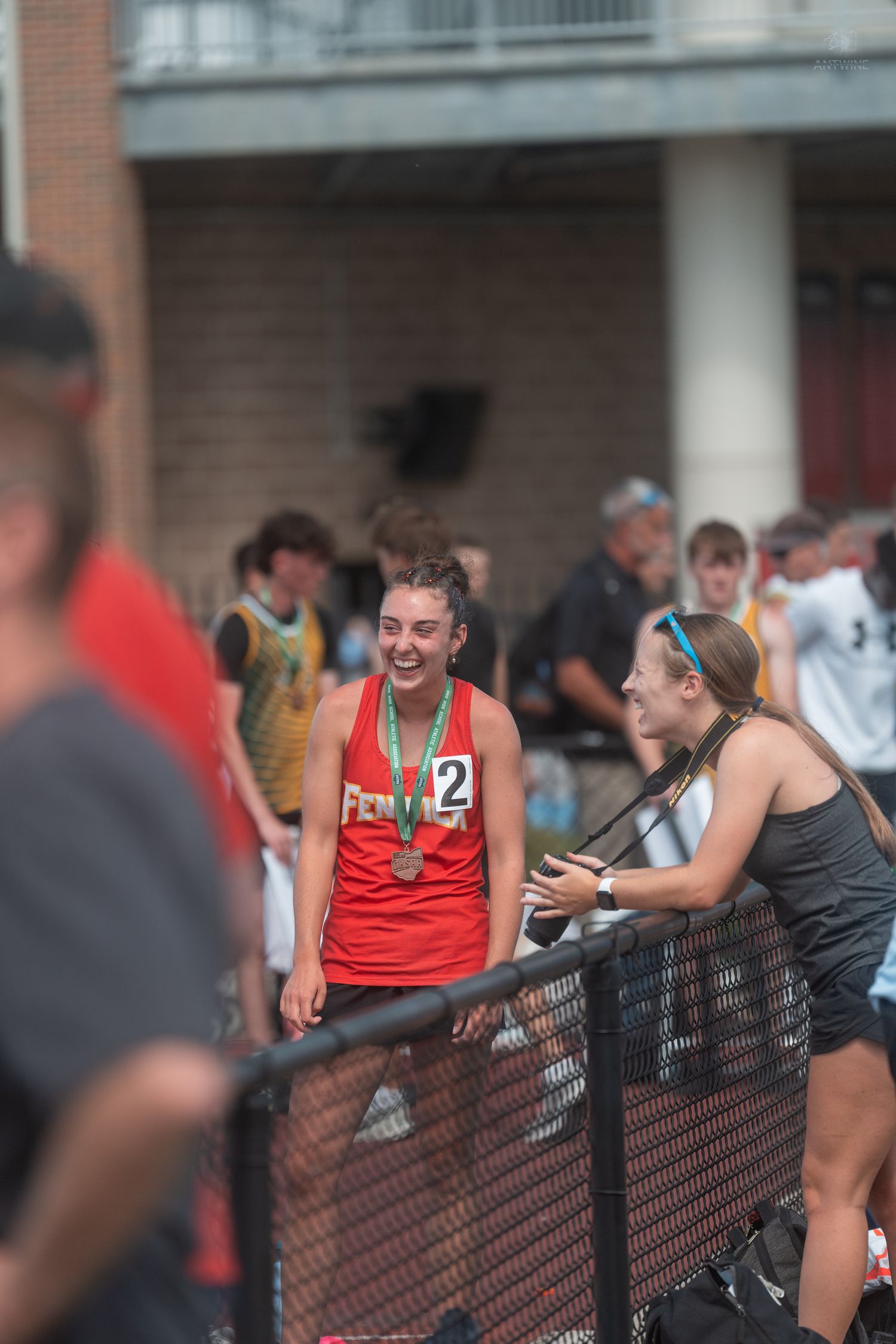 Joyful female athlete in red jersey displays medal while talking with friend at fence.