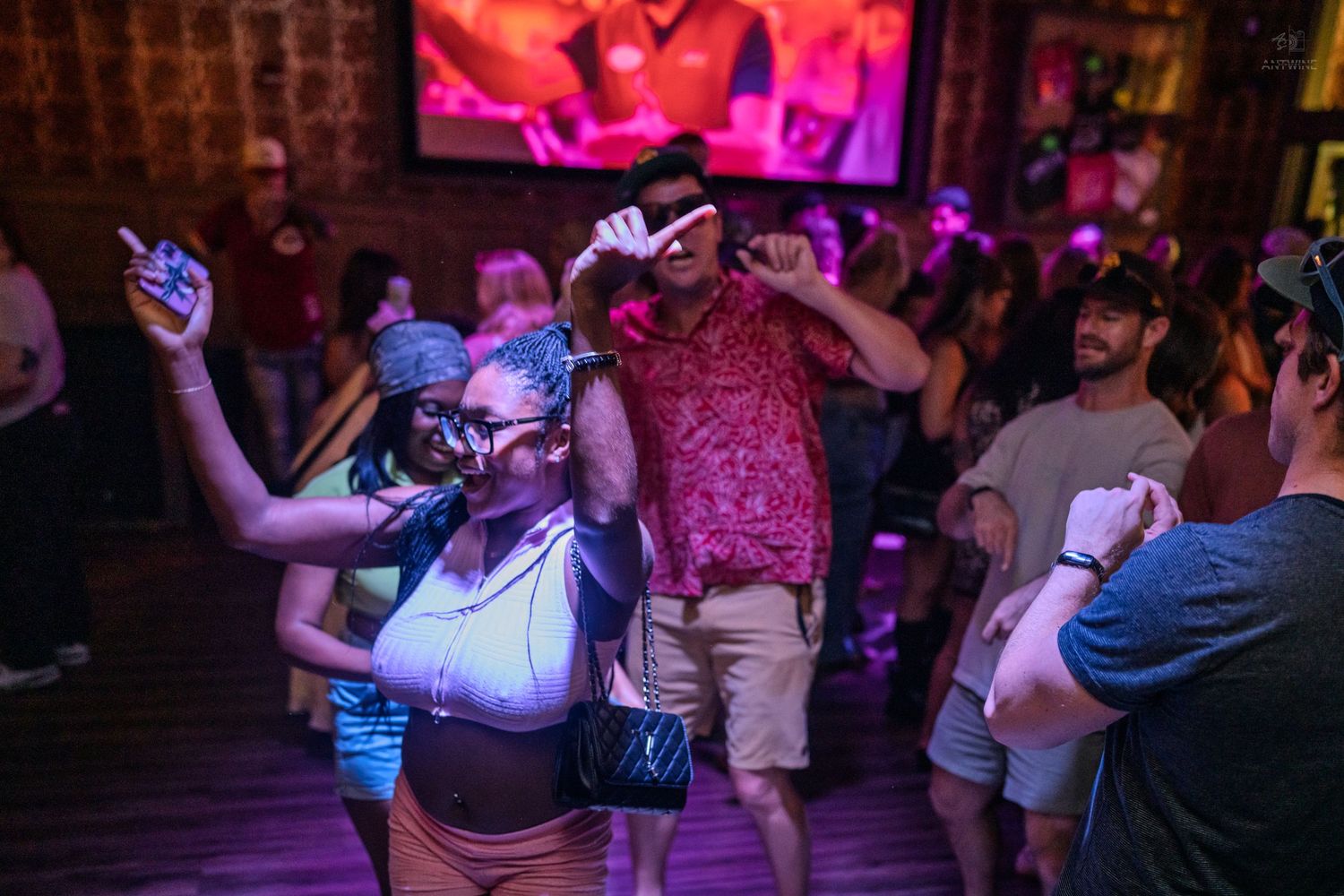 People dancing energetically inside a dimly lit bar with purple lighting and a large projection screen.