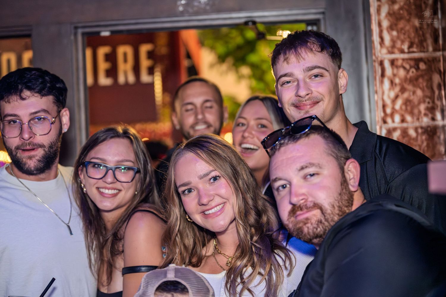 Group of seven young people smiling and posing together outside a bar at night.