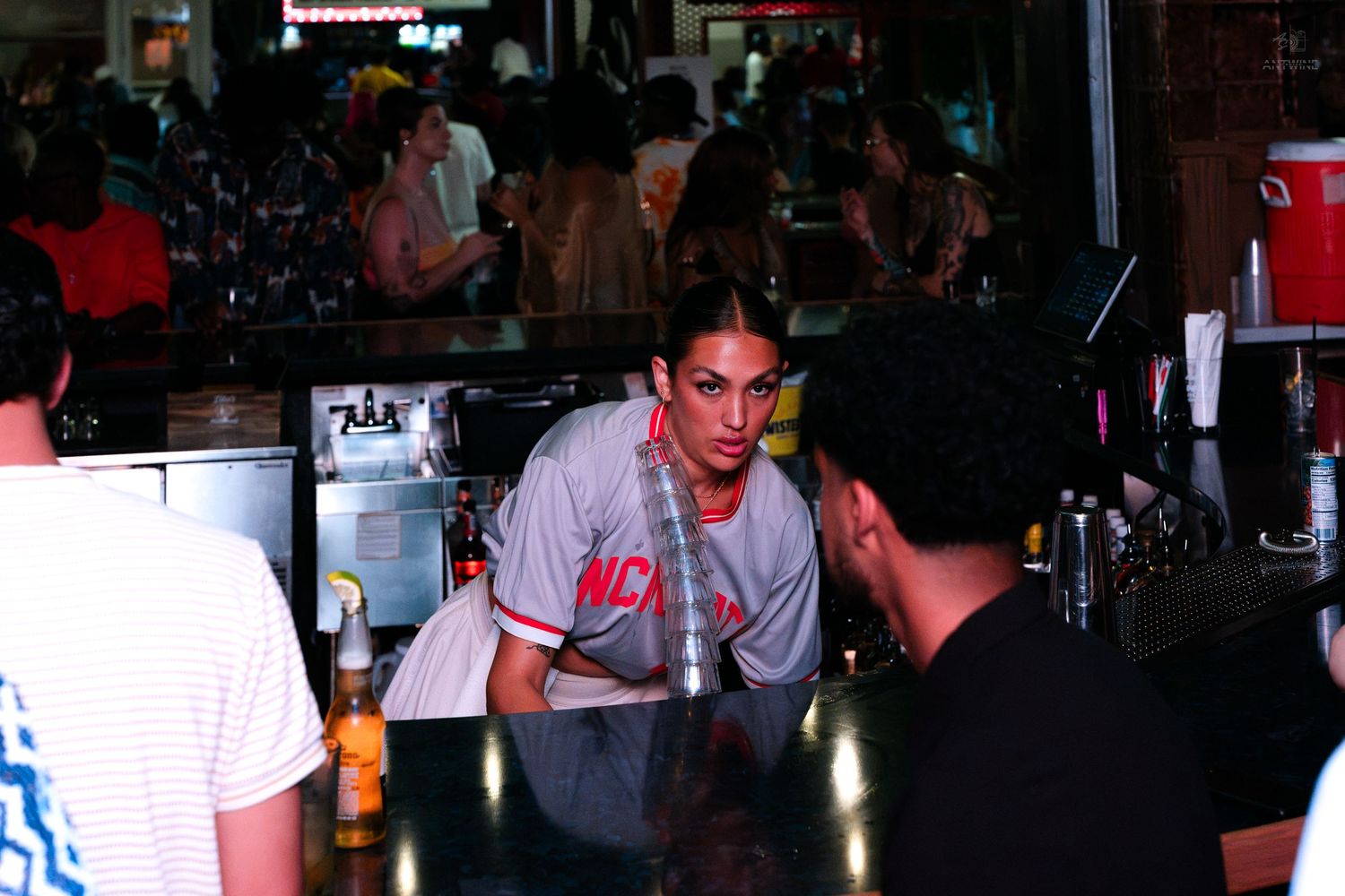 Female bartender leaning over a dark bar counter serving a customer at a busy nightlife establishment.