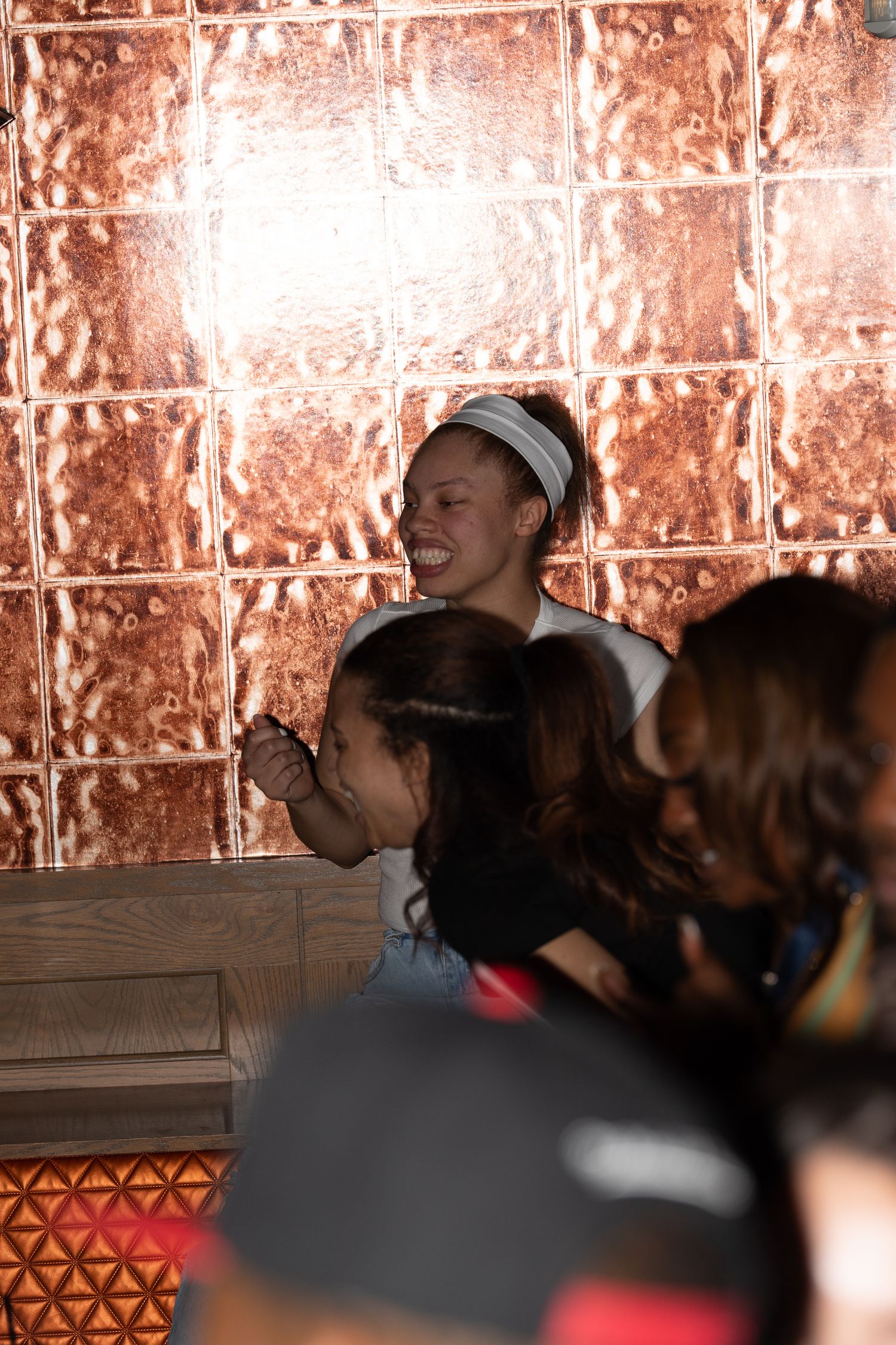 Smiling woman in white headband stands against a copper tile wall at an event.