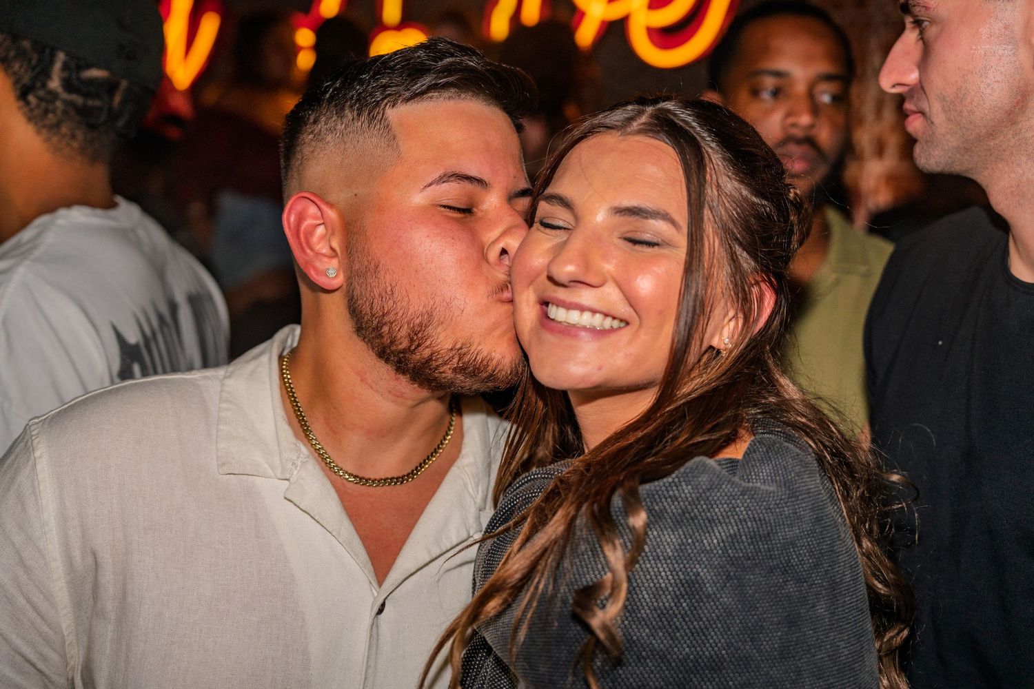 Young couple sharing a kiss at a crowded nightclub with neon lights in the background.