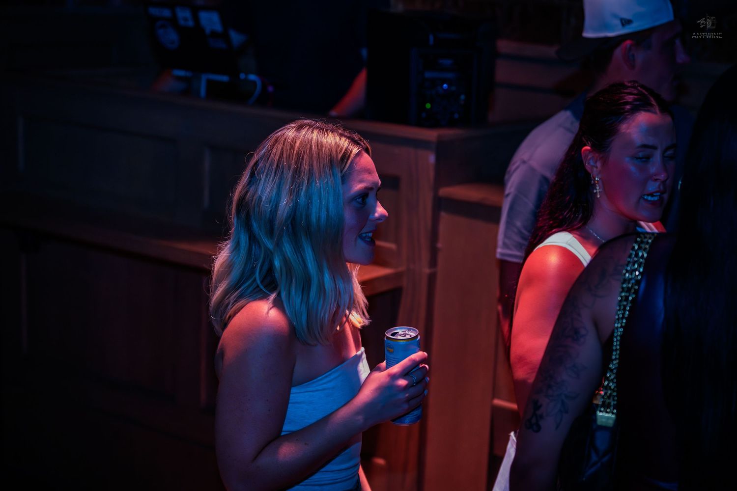 A blonde woman in a blue top holds a drink can at a dimly lit nightclub event.